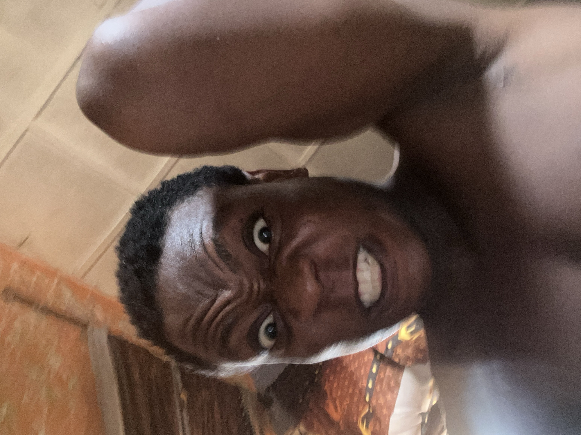 The image shows a close-up, low-angle shot of a dark-skinned person with short, dark hair. The person's face is contorted with furrowed brows and visible teeth, giving an intense or aggressive expression. A large bicep fills the upper part of the frame. The background is blurry, but suggests the edge of a wall and what appears to be a patterned object. The location is Unknown, Unknown.