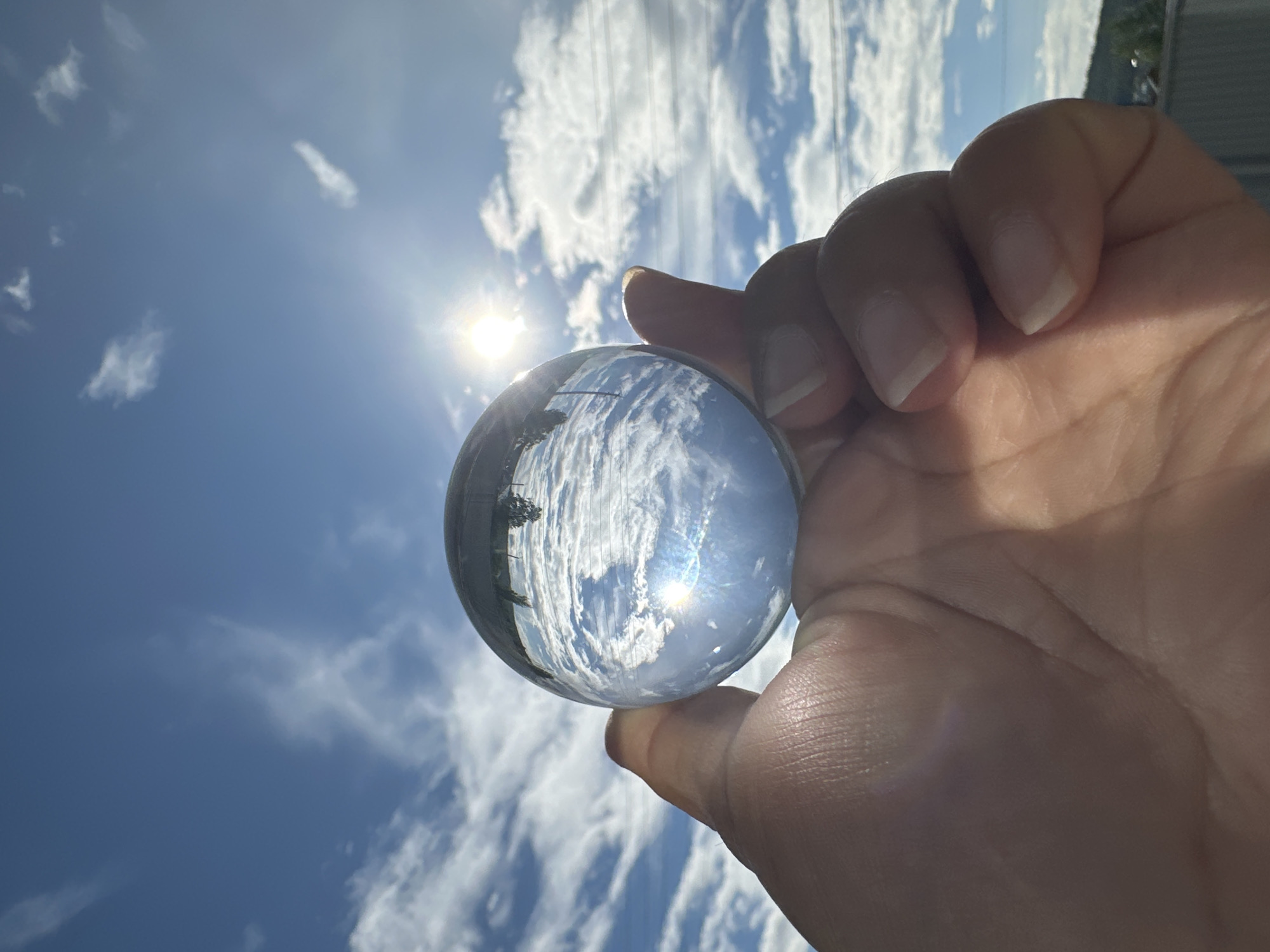 A human hand, with light skin and well-maintained nails, holds a clear spherical crystal ball upwards against a bright blue sky. The crystal ball occupies the central-left portion of the frame, held between the thumb and fingers. Inside the ball, an inverted and distorted reflection displays a vibrant blue sky with numerous white clouds, a prominent sun flare, and what appears to be inverted distant trees or structures along the top edge of the reflection. The background, outside the ball, features a vivid blue sky with scattered white cumulus clouds. The sun is intensely bright in the upper right, casting a strong lens flare. Horizontally across the mid-ground, thin power lines are visible, stretching across the scene. In the lower right background, a distant green hillside or mountain range is discernible, with the faint outline of structures or buildings beneath it. The scene is illuminated by strong, direct sunlight. The location is in the city of Unknown, Unknown.