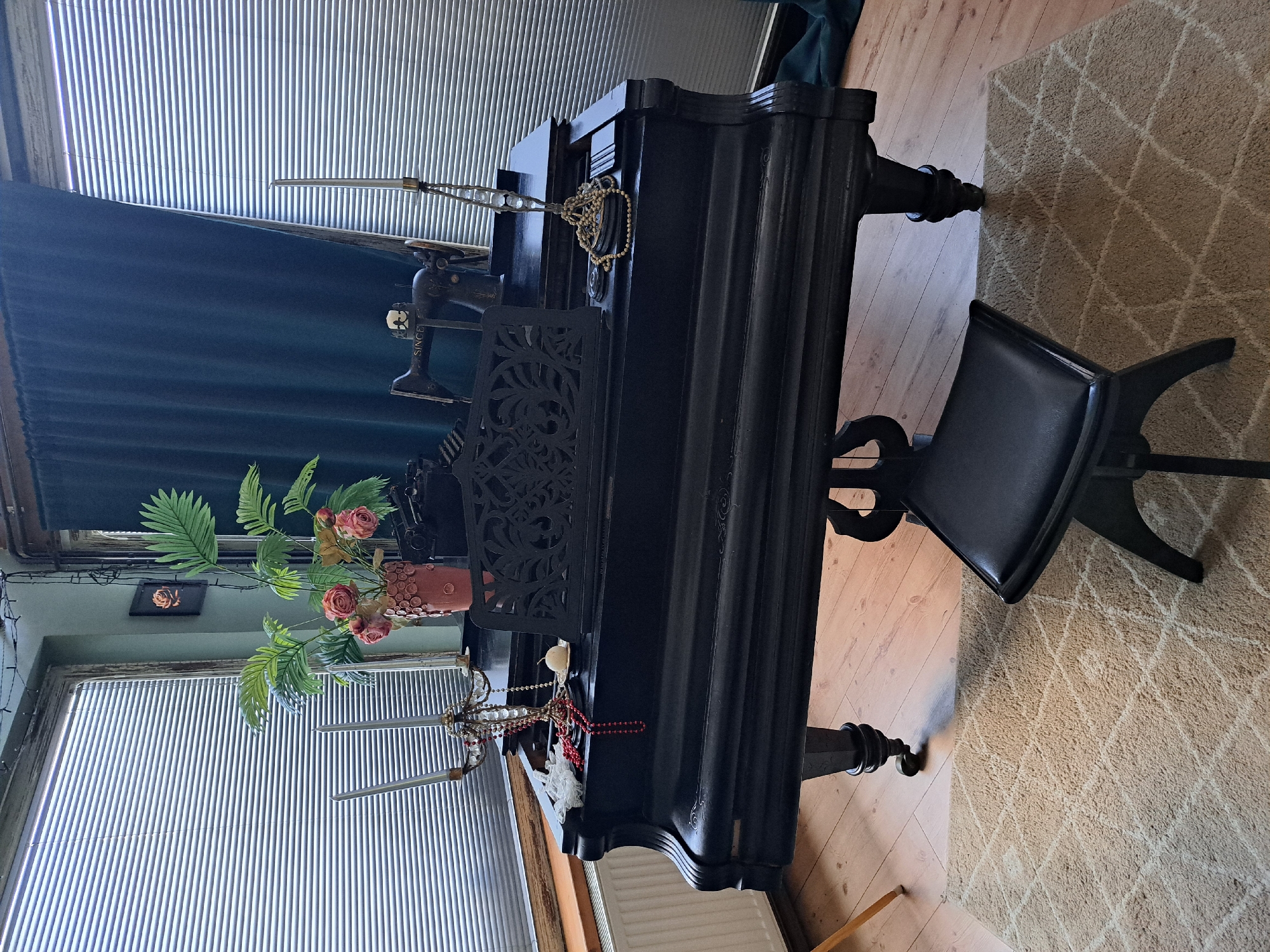 The image shows a black upright piano in a room with wooden flooring and a patterned rug. A black piano bench is positioned in front of the piano. On the piano, there is a small black sewing machine, a gold necklace, and a vase with faux flowers. To the left of the piano, there is a window with blinds and a dark blue curtain. A string of lights is visible along the top left of the window frame. Another window with blinds is visible below and to the left of the piano. The lighting suggests it is daytime, with natural light filtering through the blinds. The overall scene appears to be a domestic setting, possibly a living room or a study, decorated with various objects.