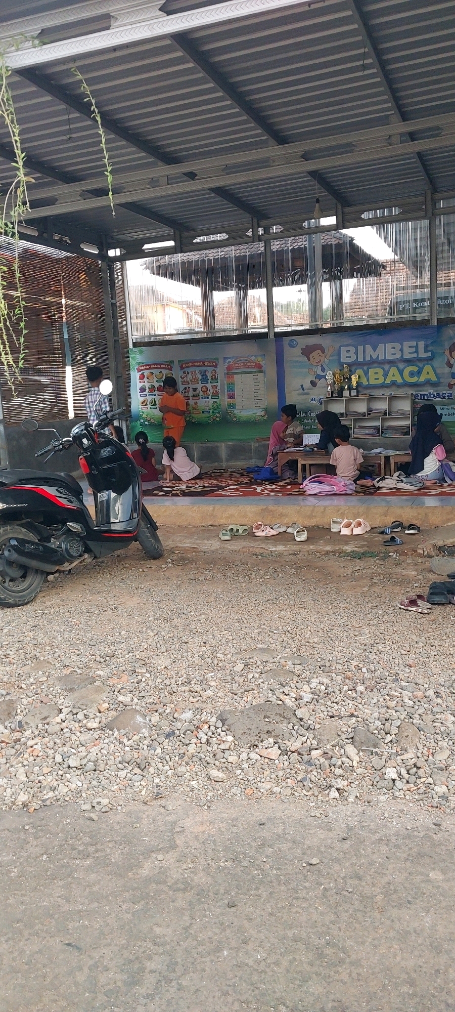 This image depicts an outdoor learning environment, likely a small, informal school or tutoring center, situated in Keling, Indonesia. The scene is set under a corrugated metal roof supported by metal beams, offering shade. In the foreground, a black scooter is parked on a gravelly ground.
The primary activity centers around a group of children and an adult engaged in what appears to be a learning session. Several children are seated on a patterned rug laid out on a concrete platform. One child, wearing an orange shirt, stands while facing the others. Another child, in a pink shirt, is seated on the rug, looking down. A woman, possibly a teacher, is seen interacting with children seated around a small table. In the background, a colorful banner with Indonesian text and images of fruits, animals, and numbers is displayed on the wall, indicating an educational theme. Shelves with books and trophies are also visible.
The environment is rustic, with bamboo blinds on one side and transparent plastic sheeting on the other side of the covered area. Shoes are scattered in front of the learning area, suggesting that participants remove their footwear before entering. The overall impression is one of a modest, community-based educational setting.