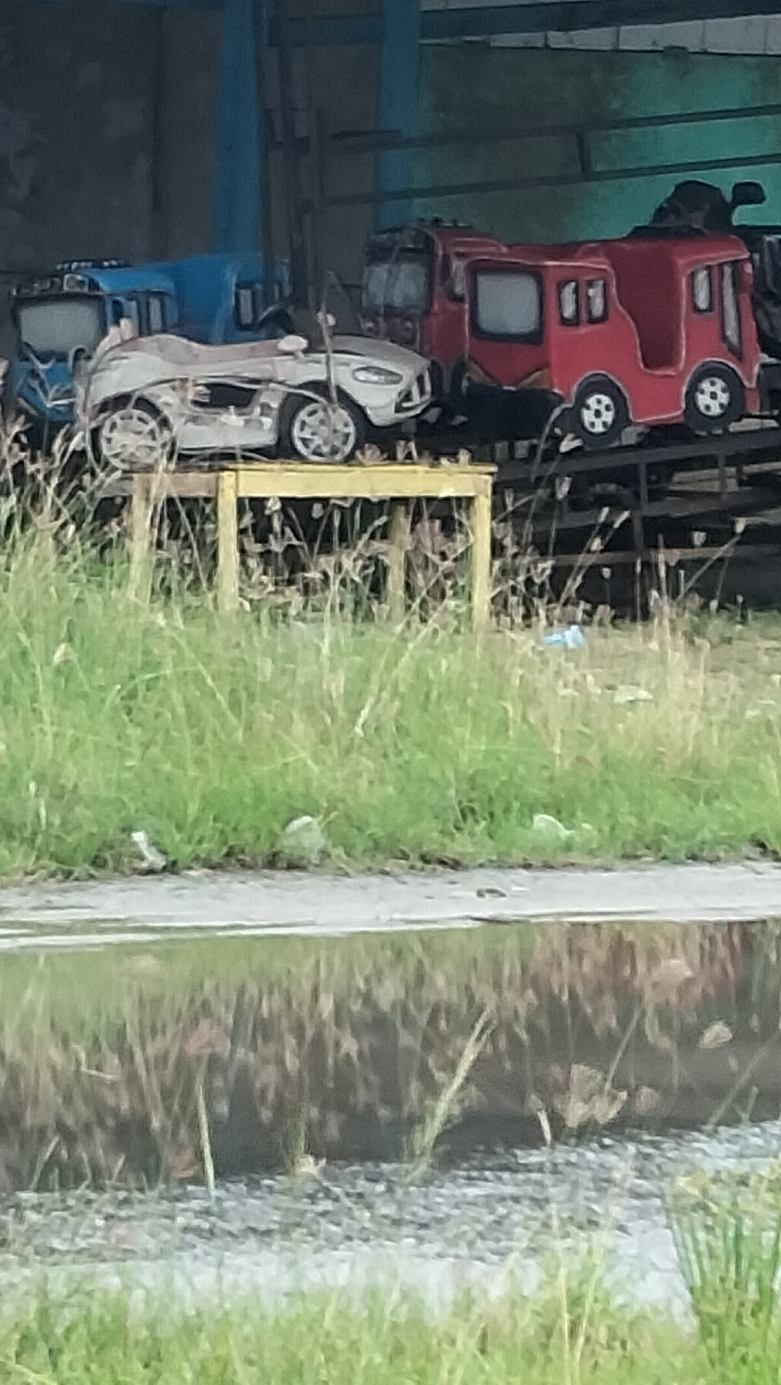 The image shows a collection of amusement park rides, likely abandoned or in disrepair, situated in an outdoor setting. In the foreground, tall green grass grows abundantly, with some dried stalks interspersed. A large puddle of murky water reflects the sky and the scene behind it, creating a distorted mirror image of the rides and the environment.
Behind the grass, several amusement park rides are visible. A white, open-topped, car-shaped ride is positioned on a yellow wooden platform. This ride appears damaged, with sections of its body missing and exposed internal components. To its left, a blue train-shaped ride is partially visible, also appearing to be in a state of disuse. To the right of the white car-shaped ride, a red bus-shaped ride is prominent. This ride has two distinct sections, resembling a bus or tram, with windows. It is also positioned on a platform or track. In the background, more structures are visible, including what appears to be a building or a covered area, with metal supports. The overall impression is one of a neglected amusement area.
While the exact location within Lhokseumawe, Indonesia, cannot be determined from the image alone, the context suggests an outdoor recreational space that has fallen into disrepair. The presence of abandoned amusement rides points towards a former attraction or a storage area for such equipment. The surrounding environment with its tall grass and a reflective puddle indicates a humid, possibly tropical climate, consistent with Indonesia. There are no people visible in the image, nor any specific actions or interactions taking place. The scene is static and conveys a sense of abandonment and decay.