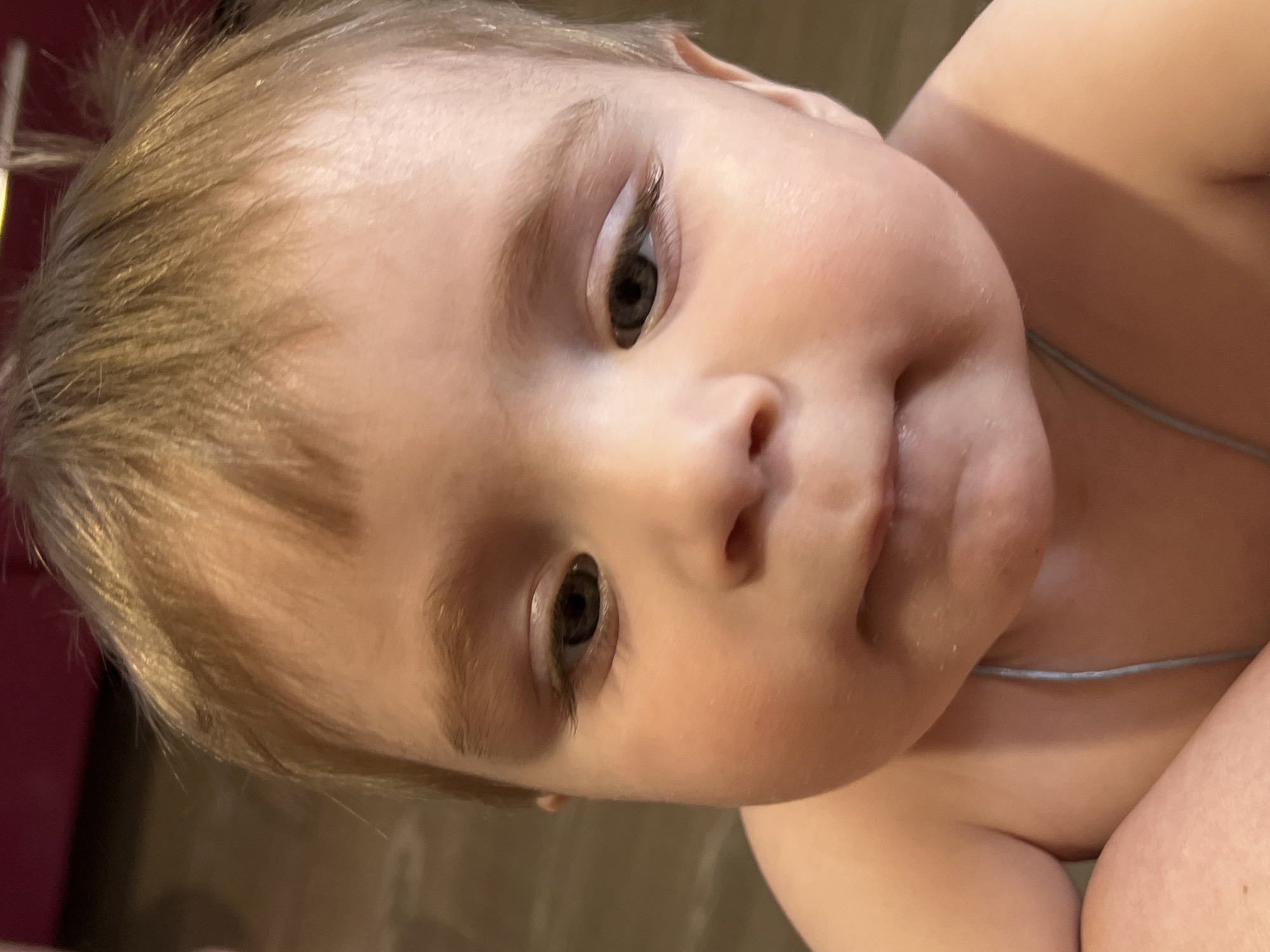 A close-up, tilted portrait captures a young baby or toddler with fair skin, light blonde or light brown hair, and blue-gray eyes. The baby's face is round with full cheeks, and the lips are slightly parted, conveying a neutral or pensive expression. A slight sheen or moisture is visible around the mouth area.
The upper body of the baby is bare, revealing smooth, soft skin. A notable detail is a very thin, light blue string or band visible around the baby's lower torso or waist. The background is softly blurred with warm, muted tones, suggesting an indoor setting. The lighting is gentle, highlighting the baby's features.
Additional cropped views emphasize details such as the fine texture of the baby's hair, the delicate eyelashes and eyebrows framing the blue-gray eyes, and the smooth skin of the body. No specific activity is depicted other than the baby looking towards the camera. The overall impression is one of an intimate and tender moment. The provided location context is Noviy, Russia. No visible text is present in the media.