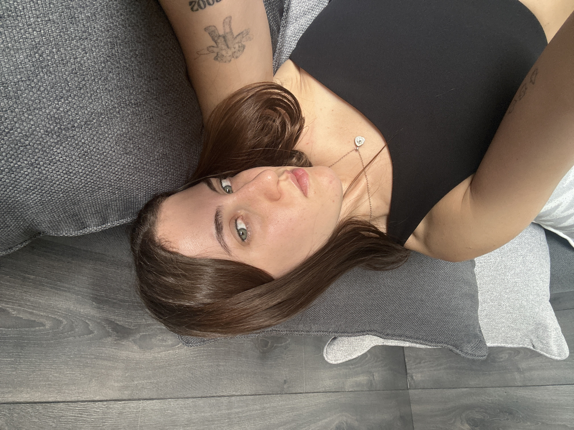 A young woman is lying down on a gray couch. She is wearing a black tank top and a silver necklace with a heart pendant. Her hair is brown and straight. Her eyes are green. She has tattoos on her left arm. The background is a gray wooden floor. The location cannot be determined.