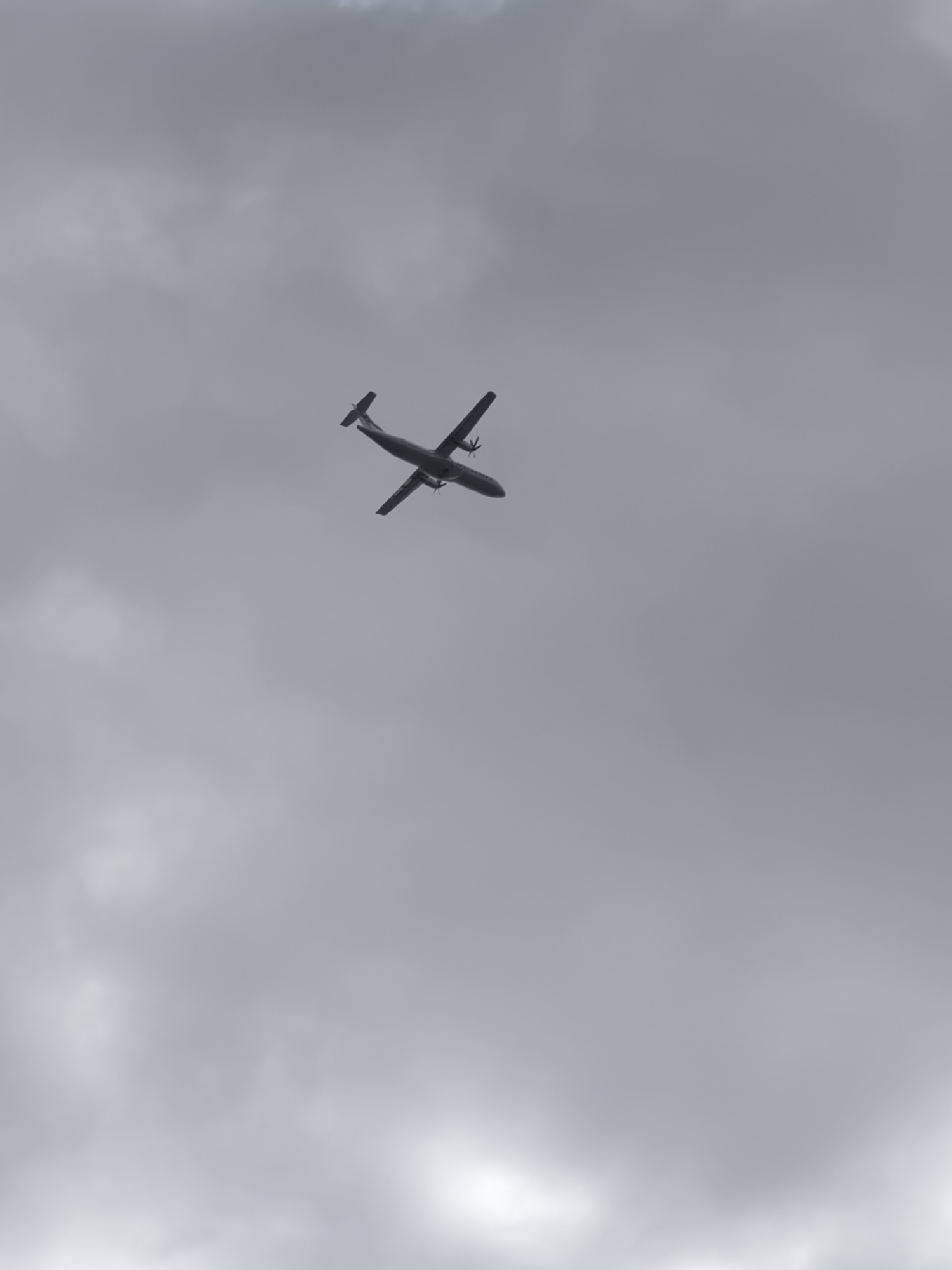 A twin-engine turboprop airplane is silhouetted against a cloudy sky, viewed from below. The aircraft's wings are angled upwards, and its propellers are visible. The image was captured at an unspecified location in Tallinn, Estonia.