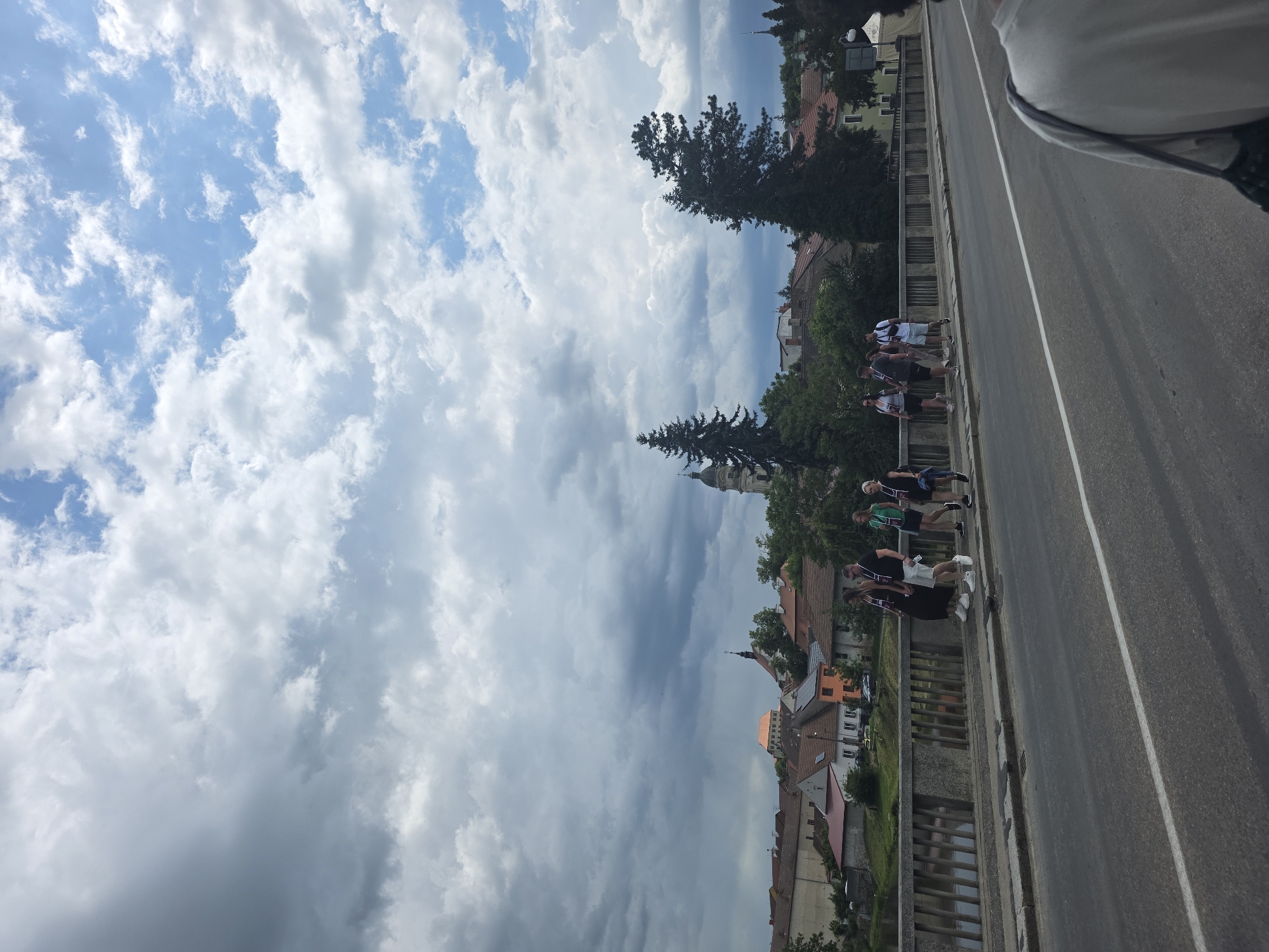 The image depicts a street scene in an urban setting. A group of people is walking along the side of a paved road. The architecture of the surrounding buildings is diverse, with visible rooftops and a church steeple. Above, a cloudy sky dominates the view. The location is the city of Unknown, Unknown.