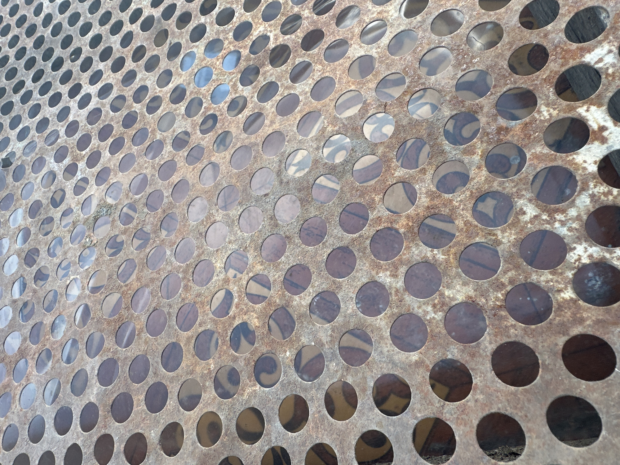 This media file captures a close-up, high-angle view of a rusted metal grate with circular perforations. The grate is tilted, creating a sense of depth and perspective. Through the holes, a patterned surface is visible, suggesting it might be a table or bench top. The pattern appears to be abstract with swirling lines and variations in color, predominantly in shades of brown, beige, and dark gray. The metal itself is weathered, exhibiting patches of rust and a textured surface. The lighting suggests it is daytime, with natural light reflecting off the metal and illuminating the scene. There are no people, animals, or specific events depicted; the focus is purely on the texture and pattern of the object. The location context of Tobyl, Kazakhstan, offers no immediate visual cues within the image itself, as the scene is generic and focuses on material detail.