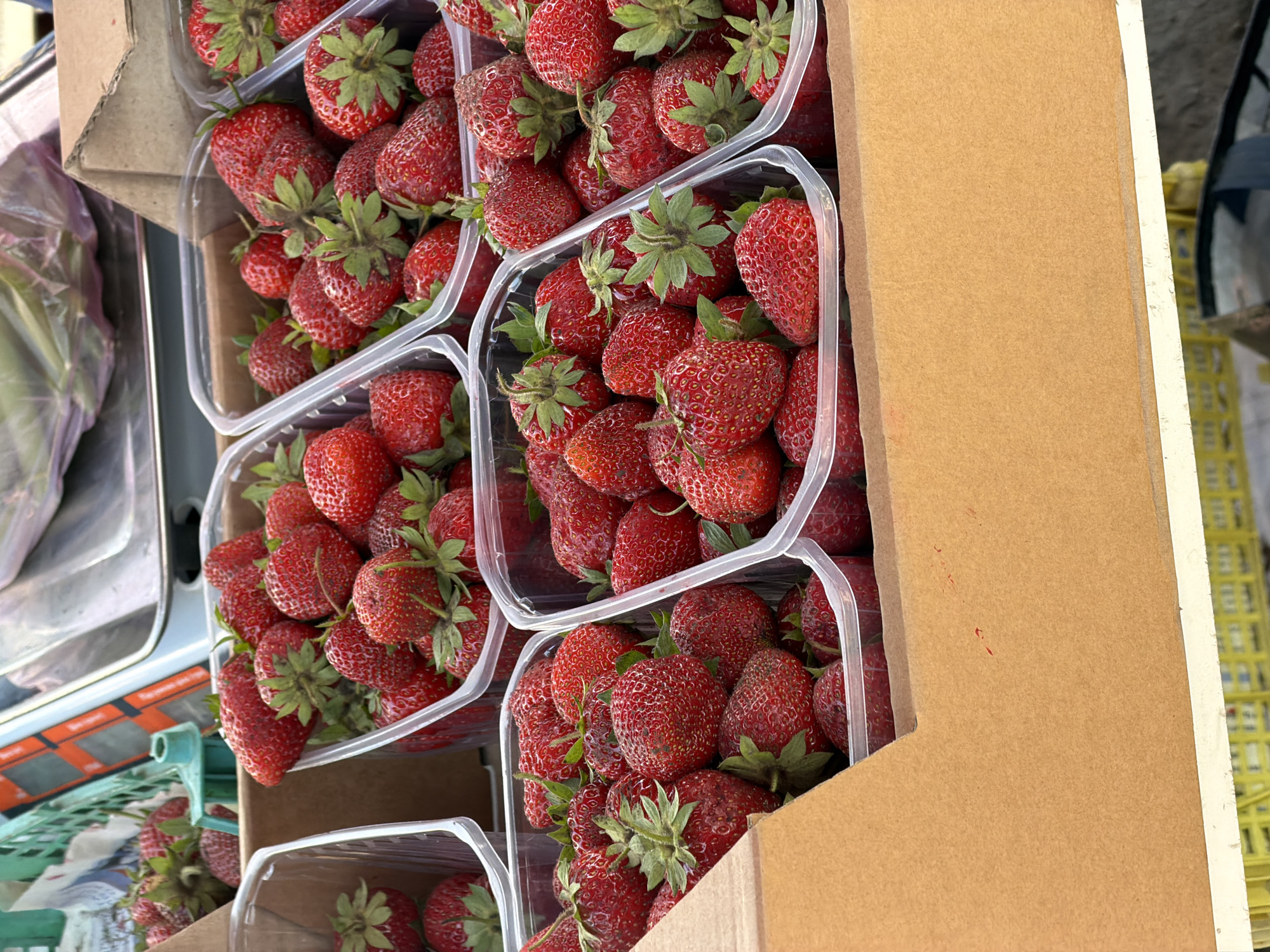 In a close-up, high-angle shot, a display of fresh strawberries is presented in clear plastic containers, nestled within a brown cardboard box. The strawberries are a vibrant red, with some showing green leaves and stems. The containers are arranged in rows, with the foreground containers being more in focus than those in the background. The lighting suggests an outdoor setting, possibly a market stall, with some background elements blurred but hinting at other produce and a cart. The overall impression is one of abundance and freshness.