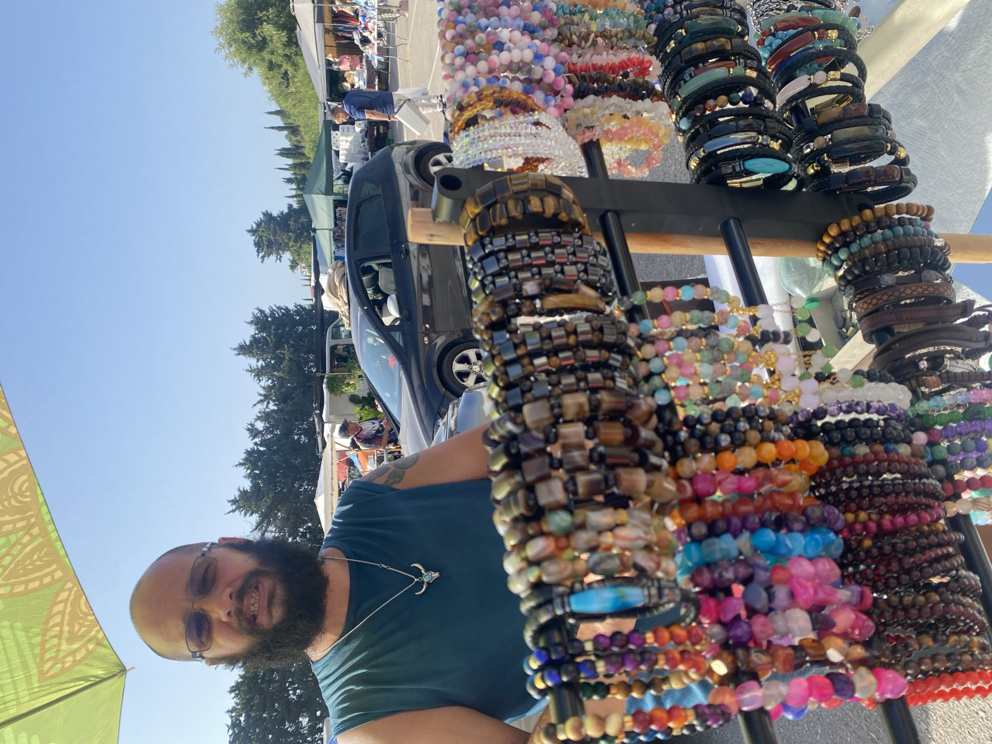 This is a medium shot, outdoor, daylight photograph taken at a flea market in Manilva, Spain. The foreground is dominated by a display rack filled with numerous colorful beaded bracelets and leather bracelets. A man, appearing to be a vendor, stands behind the display, looking directly at the camera. He is wearing a dark teal sleeveless shirt, glasses, and has a beard. He is also wearing a necklace with a bull skull pendant. Behind the vendor, a black car is parked, and other market stalls with tents and merchandise can be seen, along with trees and a clear blue sky. The overall impression is that of a busy outdoor market on a sunny day.