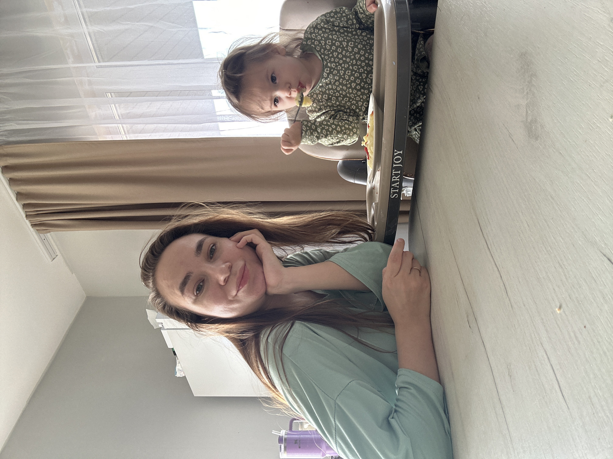 A woman and a child are depicted in an indoor setting, likely a home, captured during the daytime with natural light streaming in.
In the foreground, a woman with long, dark brown hair, wearing a relaxed light green top, is leaning on a light-colored wooden surface, possibly a table or floor, with her left hand supporting her chin. She looks directly at the camera with a gentle, relaxed expression and a faint smile. A ring is visible on her left ring finger. A purple cup or thermos is partially visible in the lower left corner of the frame.
Behind the woman and slightly to her right, a young child is seated in a dark grey high chair with a beige seat. The child, dressed in a green long-sleeved top with a light pattern, is actively eating, holding a fork to their mouth with some food visible around their lips, and looking towards the camera. The high chair has the brand name "START JOY" printed vertically on its side.
The background features a large window covered with sheer white curtains and beige blackout curtains, indicating ample natural light. The walls of the room are painted in light grey and white tones. The wooden surface, possibly a floor or table, extends across the scene and shows some scattered crumbs or food particles, suggesting a recent or ongoing meal. The overall atmosphere is casual and domestic.