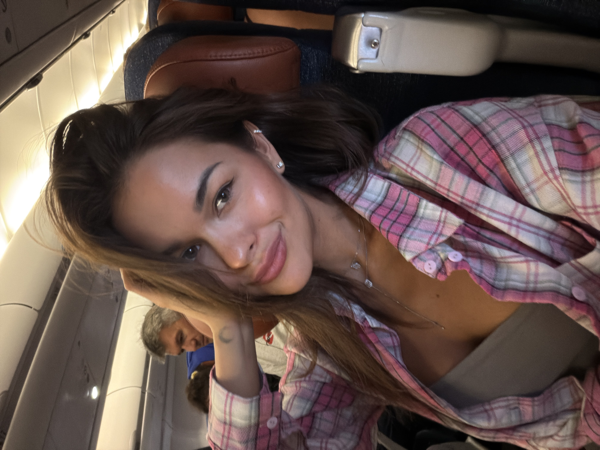 A woman is sitting in an airplane seat. She is wearing a pink and white plaid shirt over a tan-colored top and a silver necklace. The plane's interior is visible, including the seat headrest, overhead bins, and the back of another passenger’s head in the background. A man is looking at her. The image's location is unknown, as there is no information in the photo to verify it as the city of Krasnoyarsk, Russia.