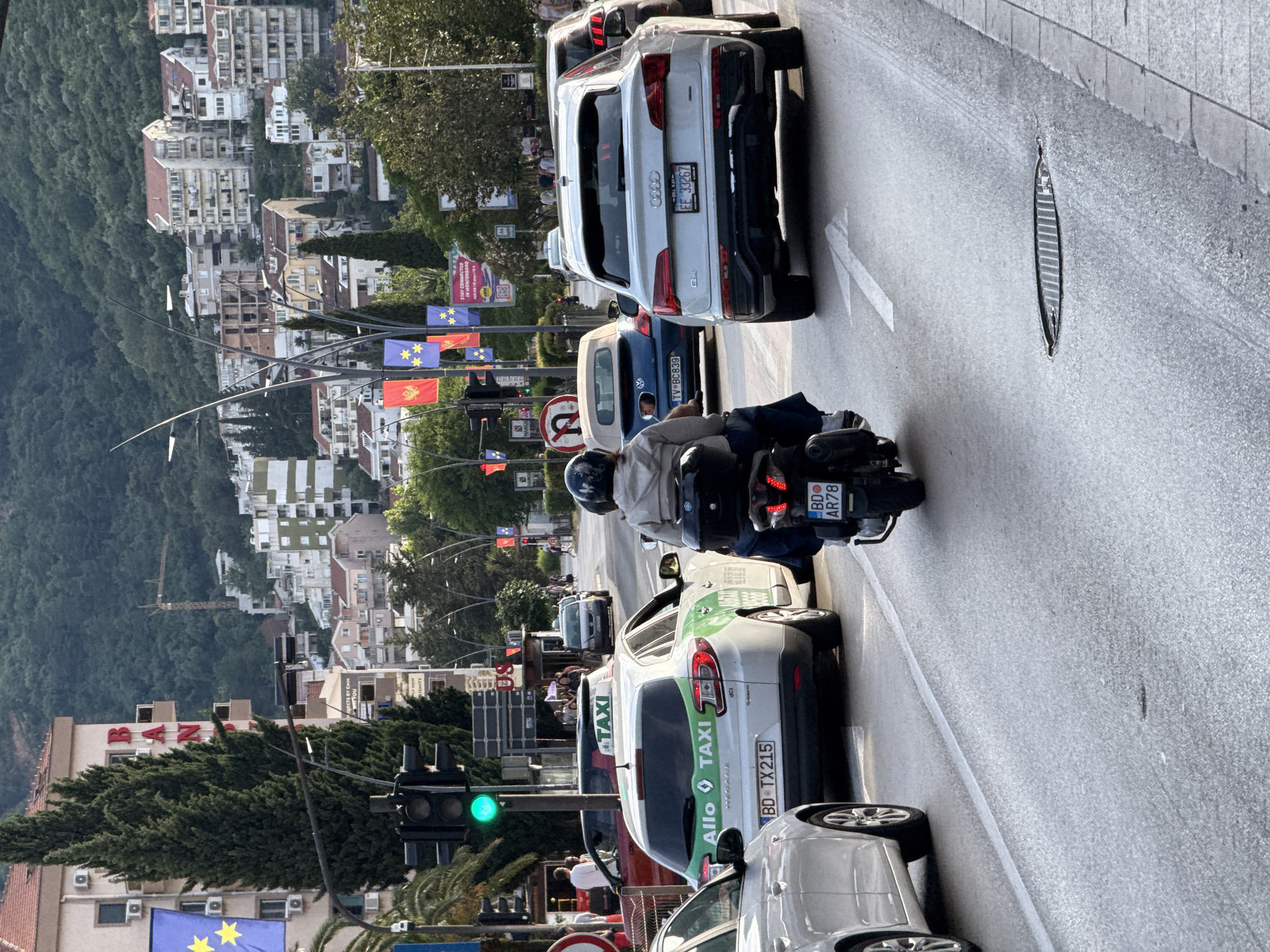 This image presents a detailed street scene in Budva, Montenegro, observed during daylight, likely late afternoon. Traffic is moving away from the viewer on a multi-lane asphalt road. In the immediate foreground, an individual wearing a light-colored jacket and a dark helmet is riding a dark scooter, displaying the Budva license plate 'BD AR78'. Directly ahead, a silver Audi Q5 with a Montenegrin license plate 'FF 33267' is present. To the left of the Audi, a white Renault Megane taxi, featuring green side stripes and 'Allo TAXI' branding, carries the Budva license plate 'BD TX215'. Further into the scene, a blue Volkswagen Beetle convertible with its top down is partially visible, bearing the Tivat license plate 'TV BC839'. A green traffic signal is illuminated on the left, and a white arrow road marking is visible on the asphalt near a manhole cover on the right.
The midground reveals a street adorned with Montenegrin national flags (red and gold) and blue flags with yellow stars, suspended above the road on decorative streetlights. A prominent sign advertising 'STAY CONNECTED IN MONTENEGRO' confirms the location. On the left, a multi-story building displays large red letters that appear to spell "BAN...". Pedestrians are visible on distant sidewalks.
The background is characterized by a steep, densely forested mountain. Numerous multi-story residential buildings with red-tiled roofs are constructed on the lower and mid-slopes of the mountain, creating an urban expansion into the natural terrain. A yellow construction crane is discernible on the upper hillside, indicating active development.