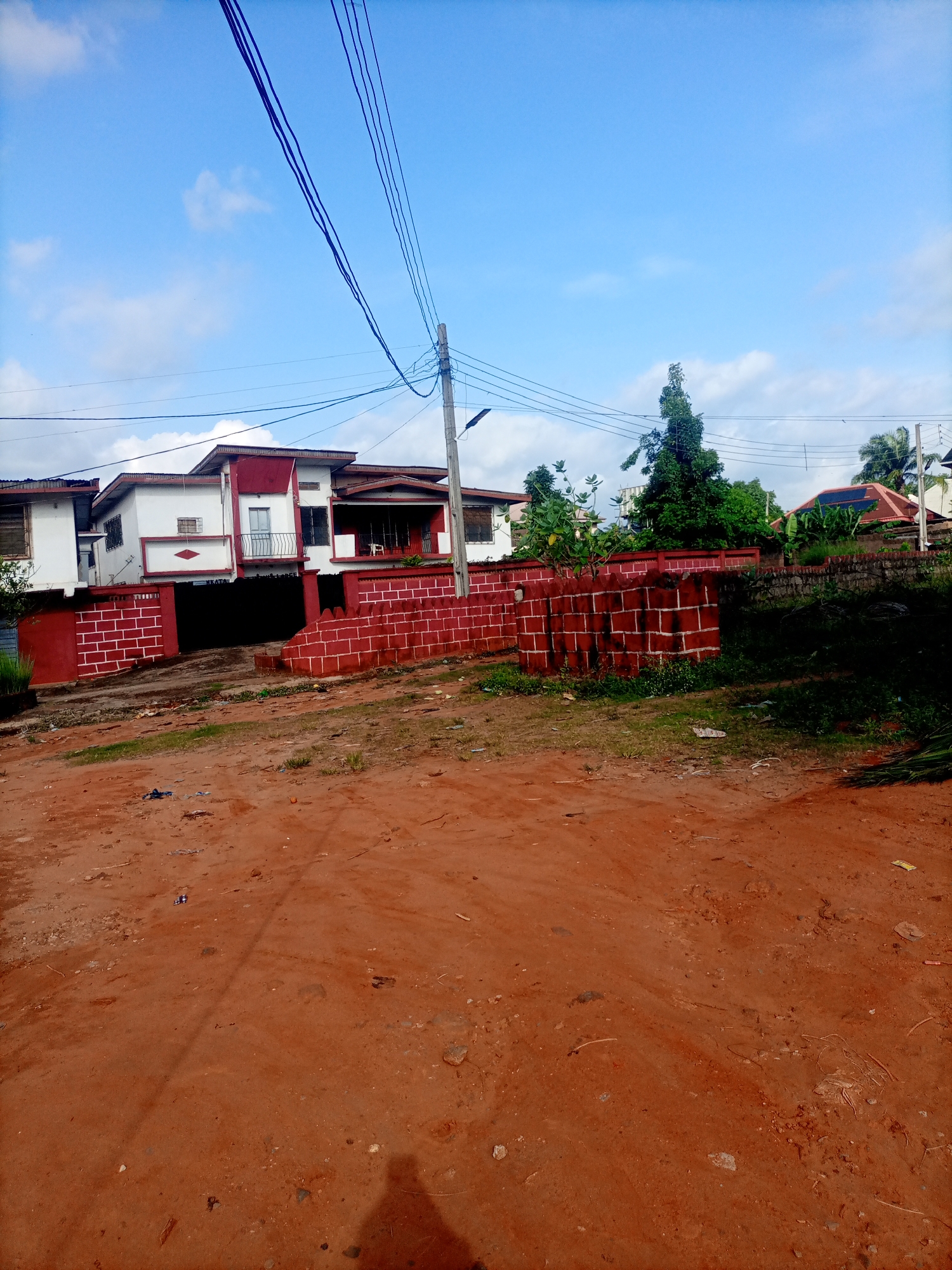 In Nkpor, Nigeria, a red dirt road leads to a two-story white and red building with a red brick wall surrounding it. The building has balconies and windows, and there are several power lines running across the sky. Green trees are visible in the background. The road is unpaved and scattered with debris, suggesting it is a less developed area. The sky is a clear blue with scattered white clouds, indicating fair weather.