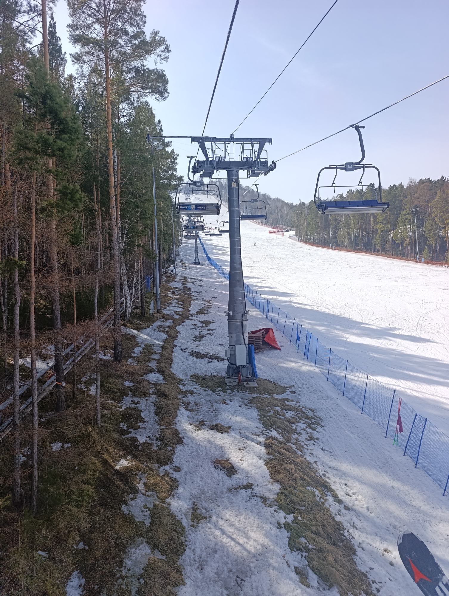 The image depicts a view from a ski lift at the Bobrovy Log Funpark, a ski resort in Krasnoyarsk, Russia. The focus is on the ski lift infrastructure, including the towers and chairs. A partially snow-covered slope is visible, with blue safety netting lining the edges. Trees are present on both sides of the slope. Patches of grass and snow are visible on the ground beneath the ski lift. A pair of skis is visible in the lower right corner of the frame. The sky is pale and overcast. No people are clearly visible on the ski slope.