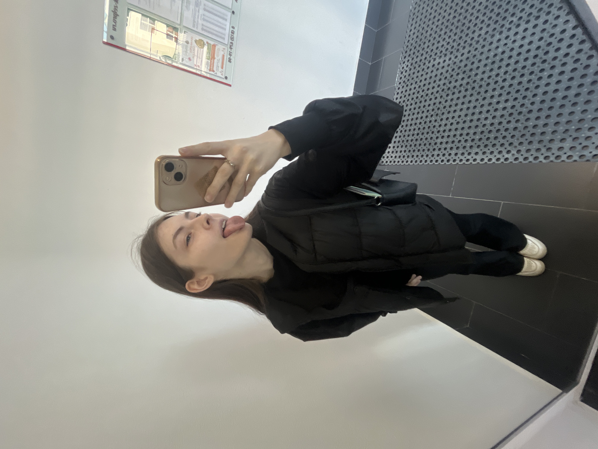 A young individual is captured in a selfie taken in a reflective surface, possibly a mirror, within an indoor setting. The person, appearing to be female with long dark hair, is shown from the chest up, extending their tongue playfully towards the camera. They are holding a light-colored smartphone with a triple-lens camera setup in their right hand, with a ring visible on one finger.
The person is dressed in a black puffer jacket over a black long-sleeved top, paired with black trousers and white sneakers. A small black shoulder bag or fanny pack is visible, slung across their body.
The setting features a plain, light-colored wall (white or off-white) on the left, which reflects the person's upper body. To the right, a dark, possibly black, tiled floor transitions into a textured, dark gray perforated metal panel that extends upwards. In the background reflection, a white information board or poster display is visible, attached to a wall. This board contains several documents or posters with text and images, and a reflection of a window showing an outdoor building and sky.
Notable details include the playful expression of the individual. Visible text on the reflected poster board, although distorted and upside down, includes what appears to be "in.org.ru" and the Russian company name "РЕСО-ГАРАНТИЯ" (RESO-GARANTIA), indicating a Russian context for the location. The overall impression is a casual, everyday moment captured indoors.