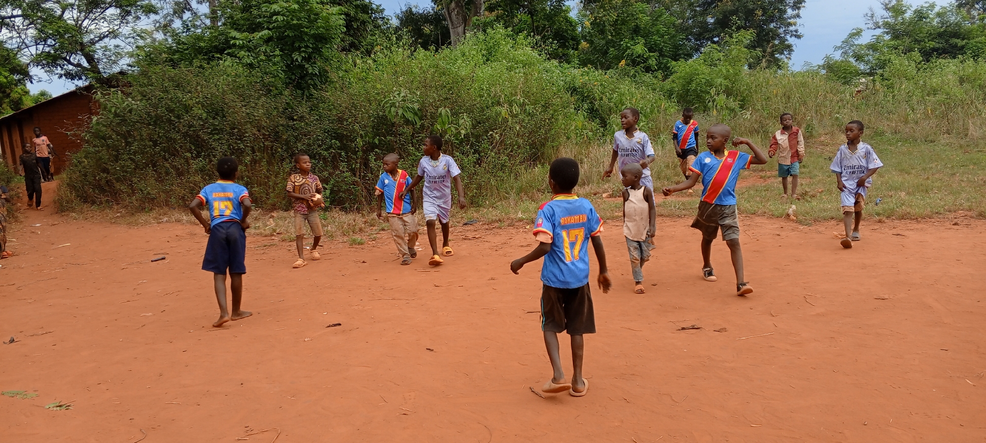 In Ngalula, Democratic Republic of the Congo, several children are playing on a dirt ground. Most of the children are barefoot and wearing sports shirts. Lush green vegetation forms a backdrop. A few people are near a building in the far left of the frame.