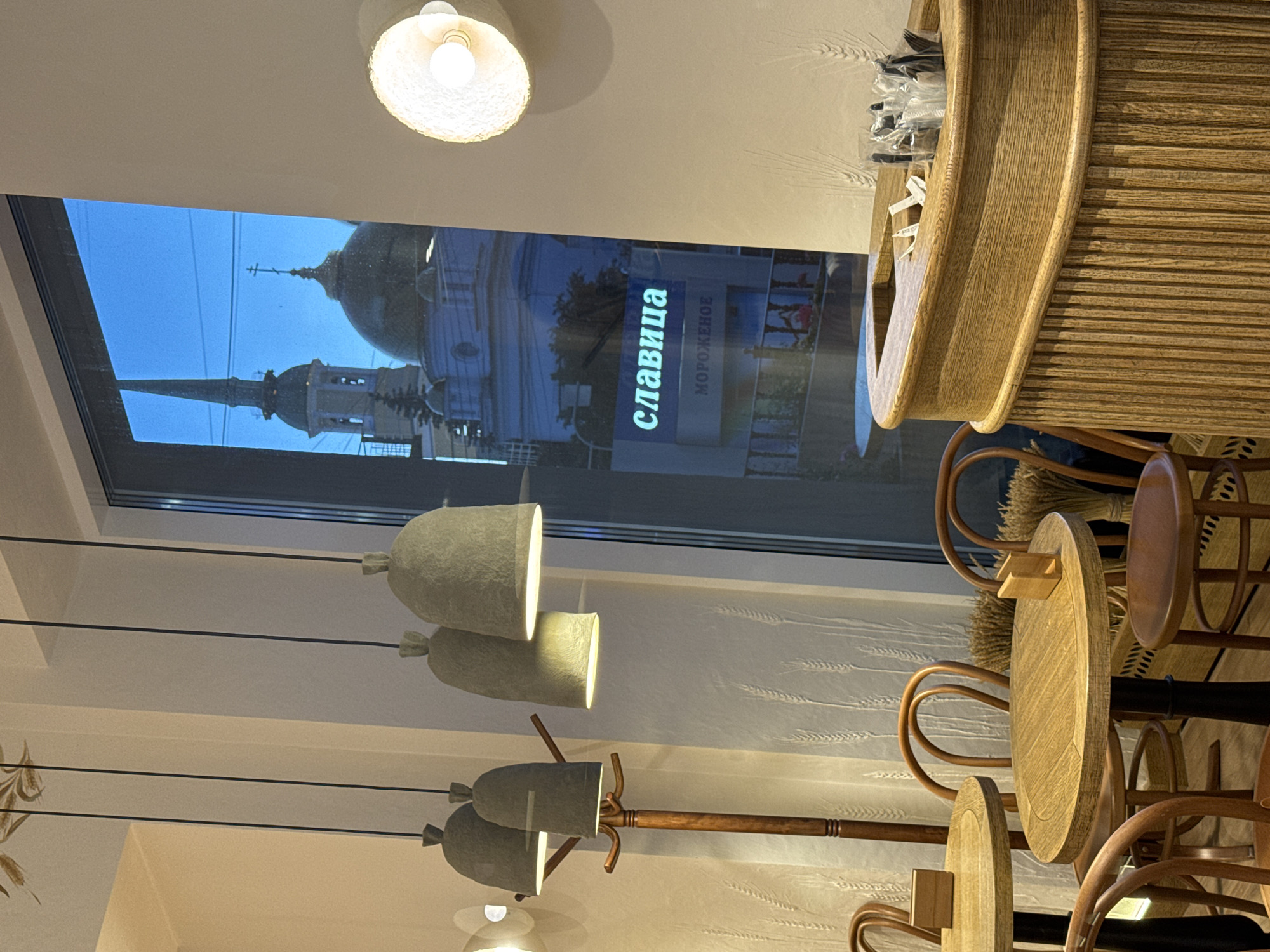 The image shows an interior view with a window reflecting an external scene. The reflection reveals the Alexander Nevsky Cathedral in Izhevsk, Russia, identifiable by its distinct domes and spire. A sign in Russian reading "славица" (Slavitsa), with the word "мороженое" (ice cream) below, is also reflected, indicating the possible location of the window. The interior features modern lighting fixtures, including pendant lamps. There are wooden bar stools and what appears to be a curved wooden counter. The overall environment suggests a contemporary café or restaurant.