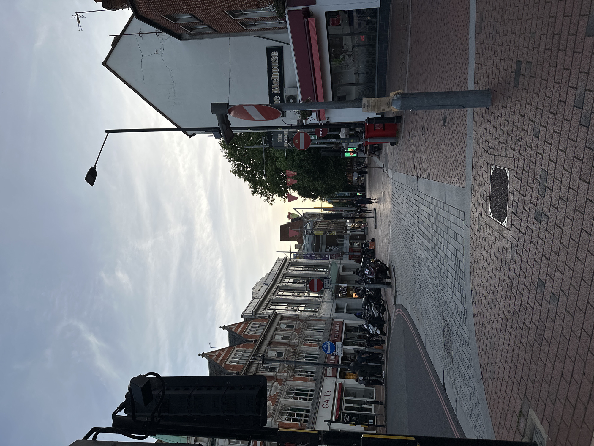 The image depicts a street scene in a city, likely during the late afternoon or early evening, as indicated by the soft light and slightly overcast sky. The perspective is from street level, looking down a paved street lined with buildings on both sides.
On the left side of the street, there are multi-story buildings with traditional architectural styles. Some of these buildings house businesses with signage visible, such as "GAIL'S." Several people are standing on the sidewalk, and a line of parked scooters and motorcycles is also present. A traffic signal pole with a black casing is prominently positioned in the foreground on the left.
The right side of the street features a white-walled building with a distinctive peaked roof and a sign that reads "The Alehouse." Adjacent to this is a shop with a red awning. A red post box and a lamppost are also visible on this side. Further down the street, more people are seen walking and sitting at outdoor tables.
The street itself is paved with interlocking bricks, and there are distinct sections of different paving materials, including a lighter gray brick pattern closer to the foreground and a darker, reddish-brown brick pattern. A road with painted white lines is visible curving away to the left. The sky above is a pale blue with wispy clouds. The overall impression is of a moderately busy urban commercial area.