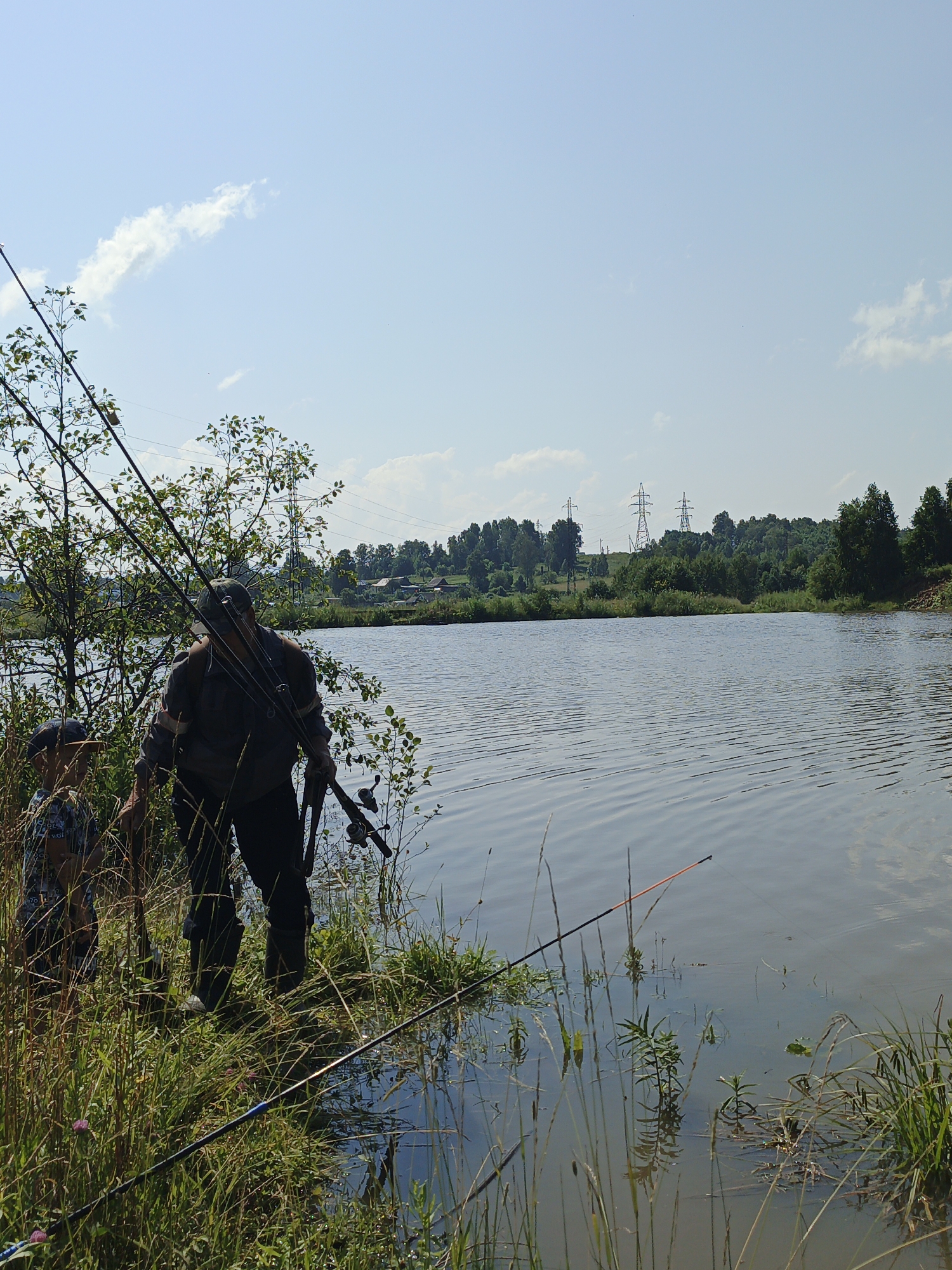 In Urtiushka, Russia, on a sunny day, two individuals, an adult and a child, stand by the edge of a calm body of water. The adult, wearing a cap and a long-sleeved shirt with a striped pattern, carries multiple fishing rods. The child, wearing a cap and a patterned shirt, stands close to the adult. The environment features lush green grass and vegetation along the water's bank, with trees and foliage further back. In the distance, across the water, a treeline is visible, with power line towers rising above the trees. The sky is a light blue with scattered white clouds. The water surface is rippled, reflecting the sky. Several fishing rods are positioned in the water and on the bank, suggesting an active fishing scene.