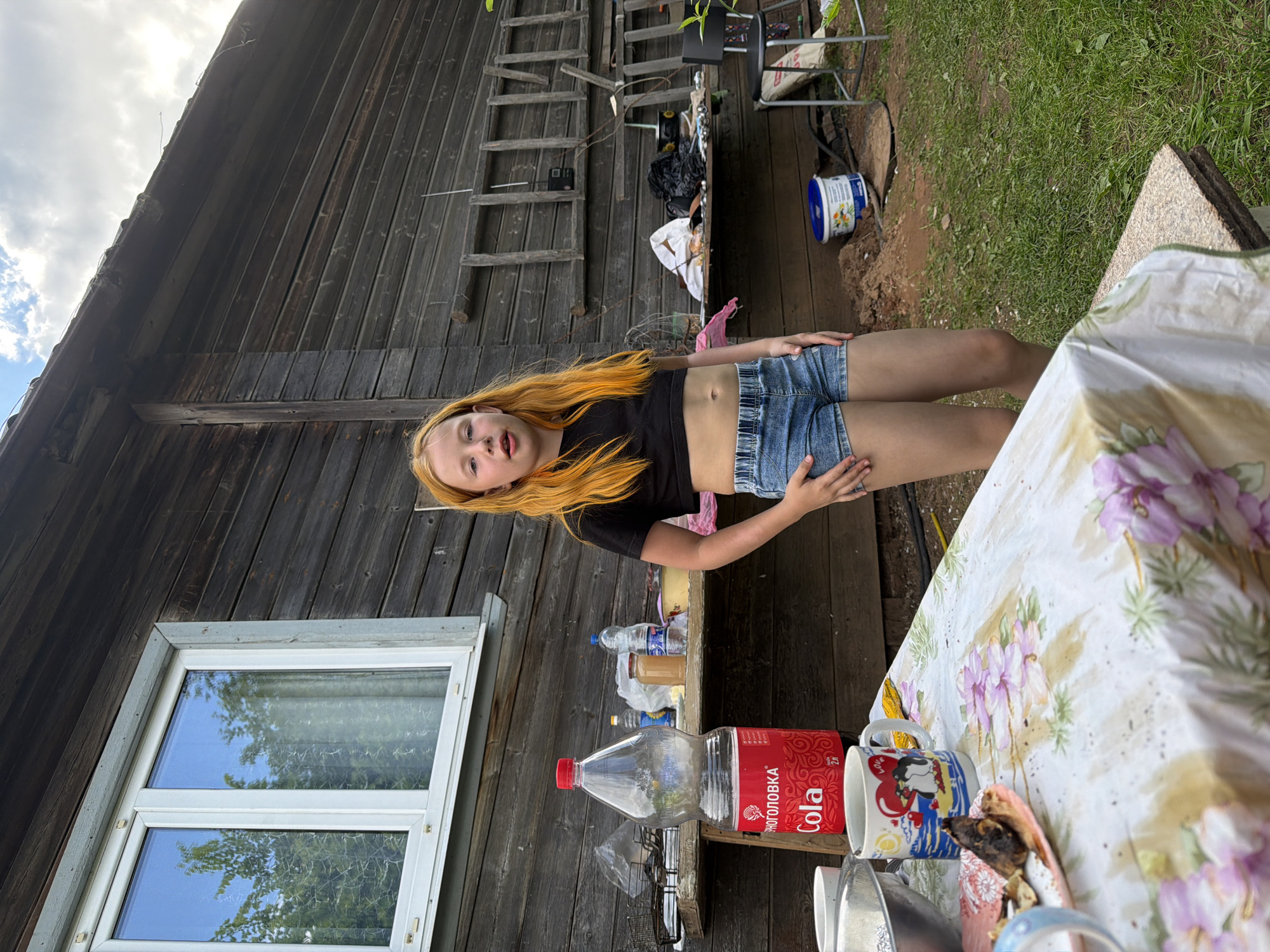 A young girl with long, bright orange hair and fair skin stands in front of a rustic wooden building. She is wearing a black crop top and denim shorts. The building appears to be a shed or small house, with weathered wooden planks and a window with white trim. A ladder leans against the side of the building, and various items, including buckets and bags, are scattered around. In the foreground, a table is set with a floral tablecloth, mugs, and a large bottle of "Cola" with Russian text. The scene suggests an outdoor setting, possibly a backyard or rural area, with green grass and dirt visible. The lighting indicates it is daytime, with the sun casting shadows. The girl's expression is neutral, and she is looking towards the camera. The overall impression is a casual, outdoor scene. The visible text on the bottle reads " Cola" and "СУПЕРКОЛО".