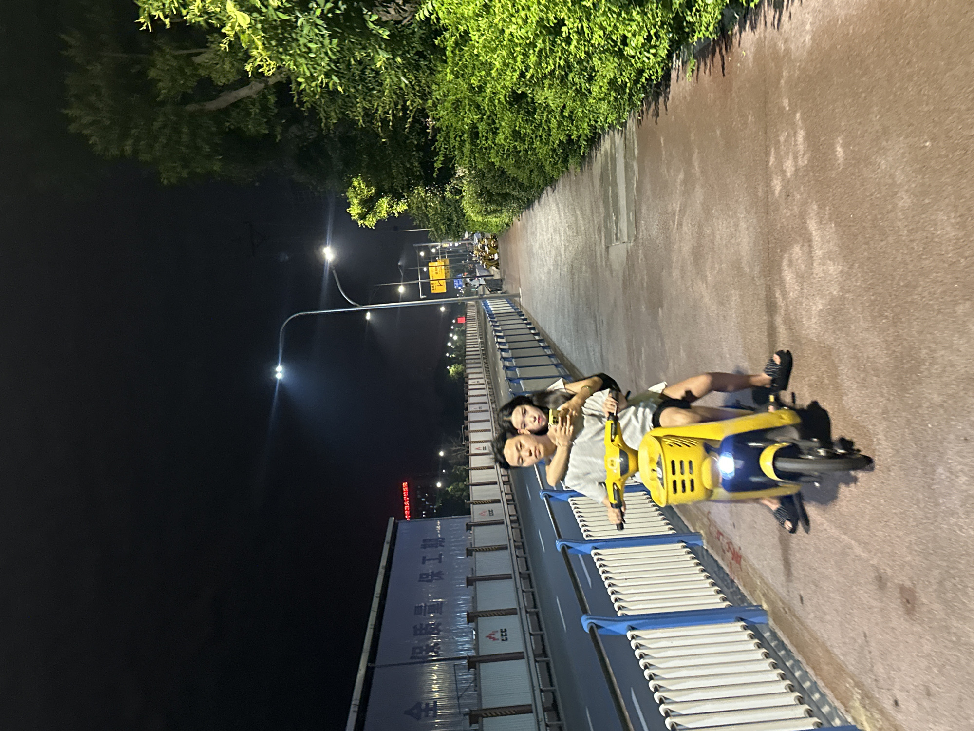 A young couple rides a yellow electric scooter at night on a sidewalk in Chongqing High-tech Zone, China. The man is driving, with his arms around the woman who is seated behind him. The woman is holding a smartphone in her right hand. They are both wearing casual clothing. The scooter has a blue front and a yellow body with a basket. The sidewalk is paved with reddish-brown material and is lined with a white and blue metal fence. Beyond the fence is a road with streetlights and a large building with signs. Lush green trees and bushes are visible on the right side of the image. The sky is dark, indicating it is nighttime.