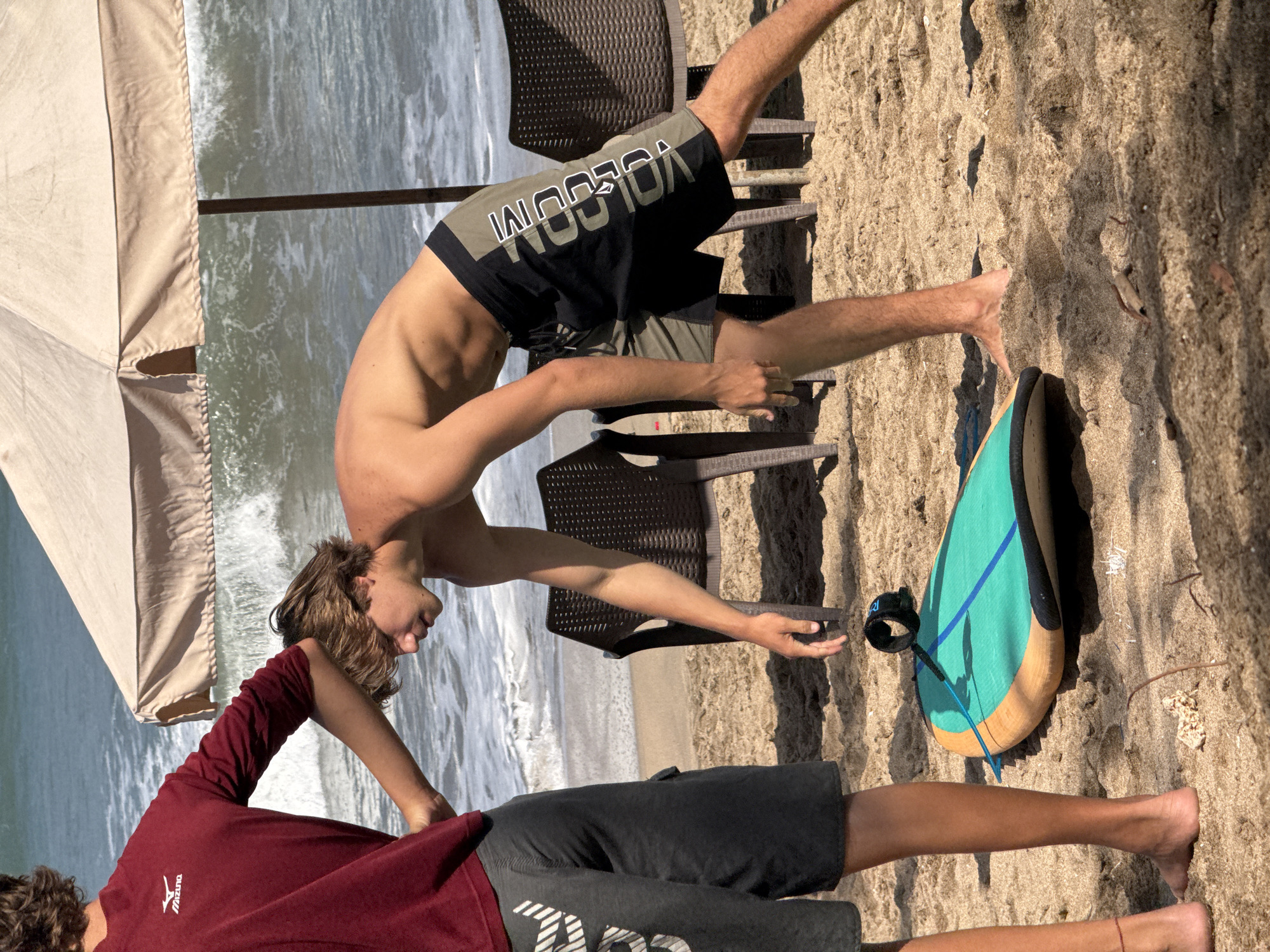 Two young men are on a sandy beach in Kuta, Indonesia, near the ocean. The man on the left is wearing a maroon long-sleeved shirt and dark gray shorts. He has his arm extended, resting it on the shoulder of the man on the right. The man on the right is shirtless, wearing black and olive green shorts with "VOLCOM" written vertically in white letters on the side. He is bending over, reaching towards the sand. In the foreground, a turquoise surfboard with orange trim is lying on the sand, attached to a black leash. In the background, waves are crashing on the shore, and a large tan beach umbrella is partially visible, along with two dark brown plastic chairs. The lighting suggests it is a sunny day.