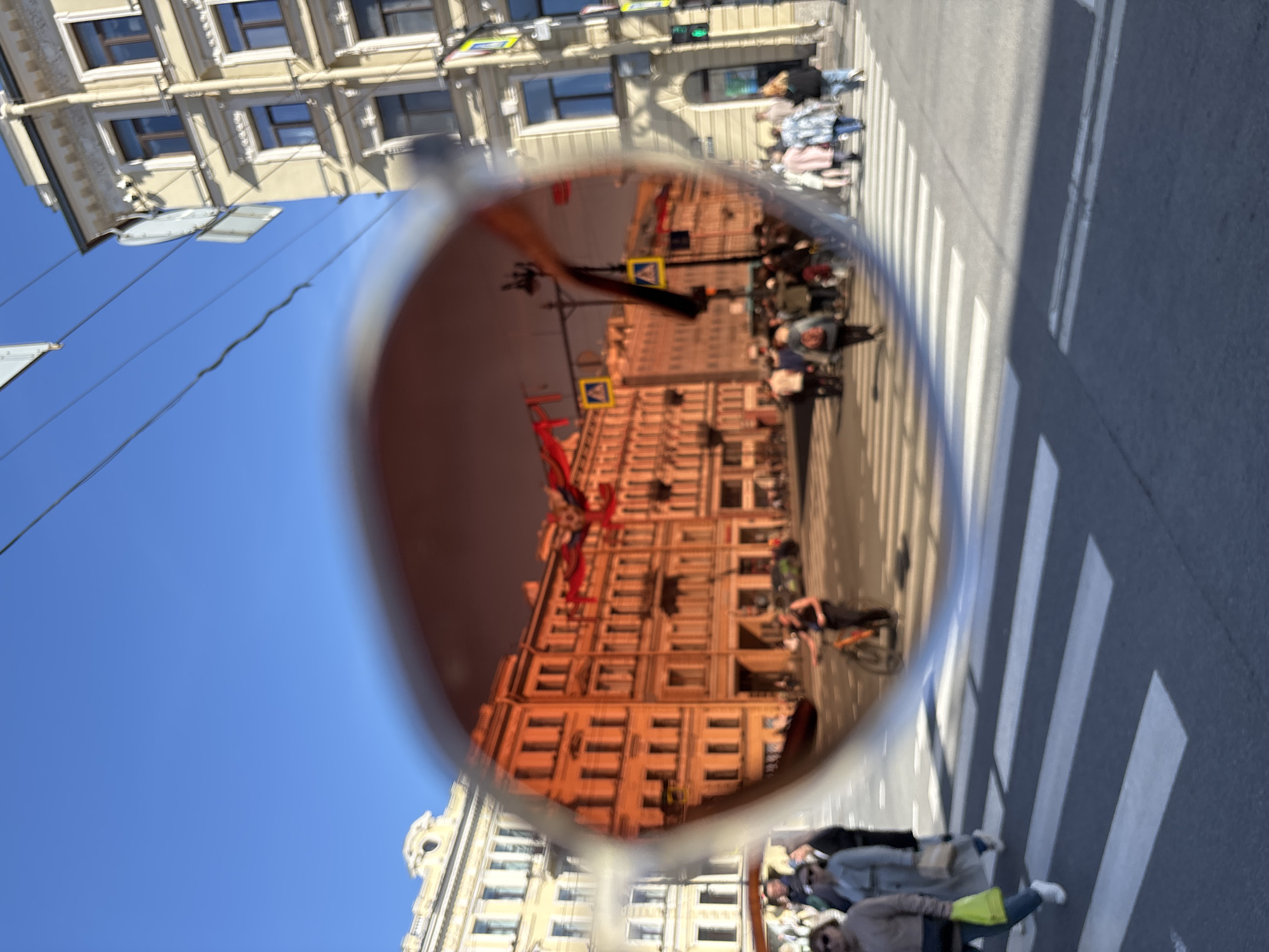 The image depicts a view of a street scene in Saint Petersburg, Russia, reflected in the lens of a pair of sunglasses. The sunglasses are held up against a background of the sky and a building's facade. The reflection shows a crosswalk with pedestrians, cyclists, and other individuals. In the background, a building is visible, along with signage and street furniture. A red decoration with streamer elements on the building is visible. The reflection suggests a sunny day. The image is vertically oriented.