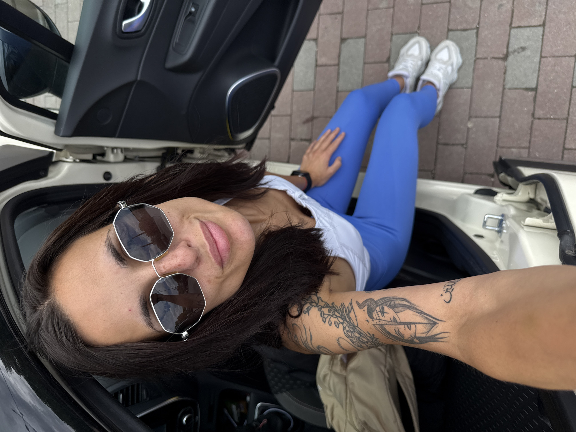 A woman with brown hair and sunglasses lies in the open trunk of a black and white car. She wears a white top and blue leggings, with white sneakers visible outside the car on a brick surface. The woman has tattoos on her arm. The photo appears to be taken from above. The location is potentially Pleven, Bulgaria, but this cannot be confirmed from the image.