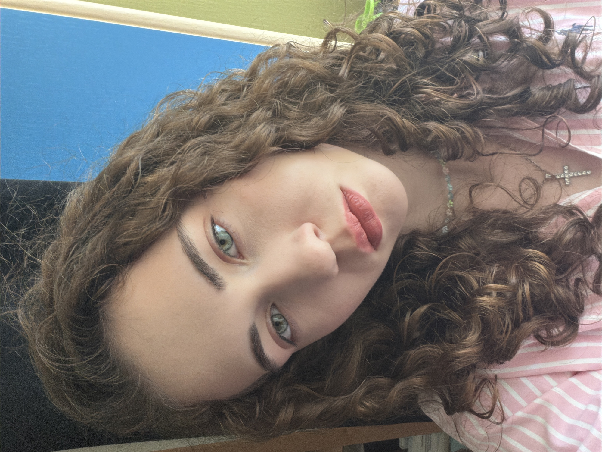 The image is a close-up, slightly angled portrait of a young woman with long, curly brown hair. She has striking green eyes and is wearing pink and white striped clothing and a delicate beaded necklace with a cross pendant. The background is blurred, featuring a vertical element that is bright blue on the left and transitions to a lighter green on the right. There is no discernible information or context to suggest this image is located in Zavodoukovsk, Russia.