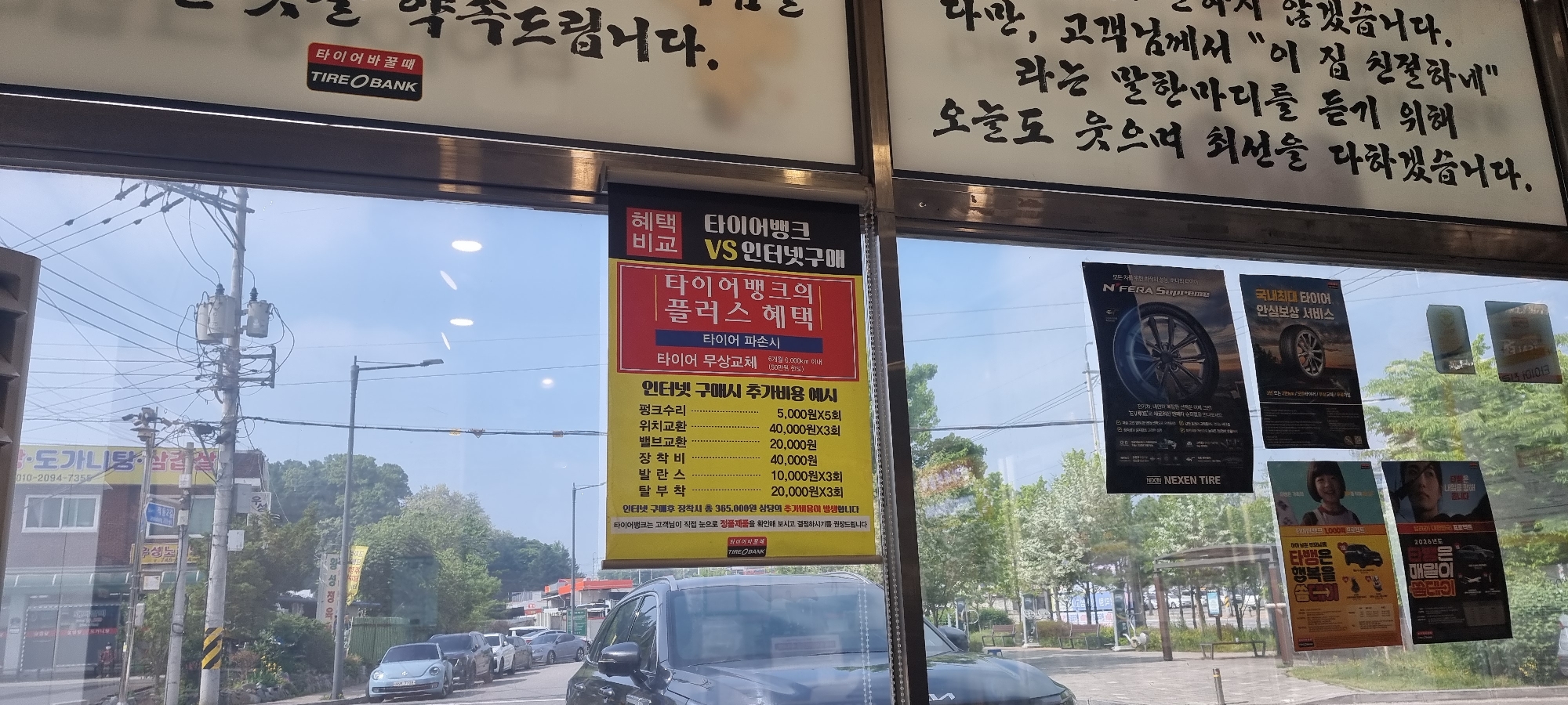 The image shows the interior view of a Tire Bank branch, likely located in Wonju-si, South Korea, as suggested by the OCR text. The view is captured through the window of the shop, which displays various posters and information about tire services. On the window, there are posters advertising tire brands, specifically N'FERA Supreme and Nexen Tire. Additionally, there's a sign comparing the benefits of purchasing tires at Tire Bank versus online, detailing potential additional costs associated with internet purchases. The text in Korean advertises the tire services offered by the Tire Bank, including free tire replacement under certain conditions. The image is framed by signage and handwritten text above the window, expressing a commitment to providing excellent customer service.
Outside, the view shows a street scene with several parked cars, including a dark-colored Kia, and a row of trees. There is also a utility pole with transformers visible. In the background, there are buildings and other structures that suggest an urban or suburban setting. The overall environment appears to be a typical day in a commercial area of a South Korean city.