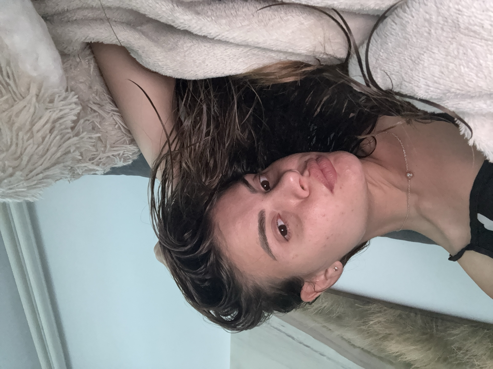 The image shows a young woman lying down, looking directly at the camera. Her head and upper body are visible, partially covered by a light-colored, fluffy blanket or bedding. Her dark brown, wavy hair is spread out around her head and appears wet or damp, with some strands clumping together.
She has wide, dark eyes and a slightly pouting or serious expression. A thin, silver chain necklace with a small, round pendant is visible on her neck, and a small silver stud earring is in her right earlobe. A dark garment, possibly a tank top, can be seen on her right shoulder. The background is a plain light blue or grey wall, indicating an indoor setting, likely a bedroom. The time of day and weather are not discernible. No text is visible.