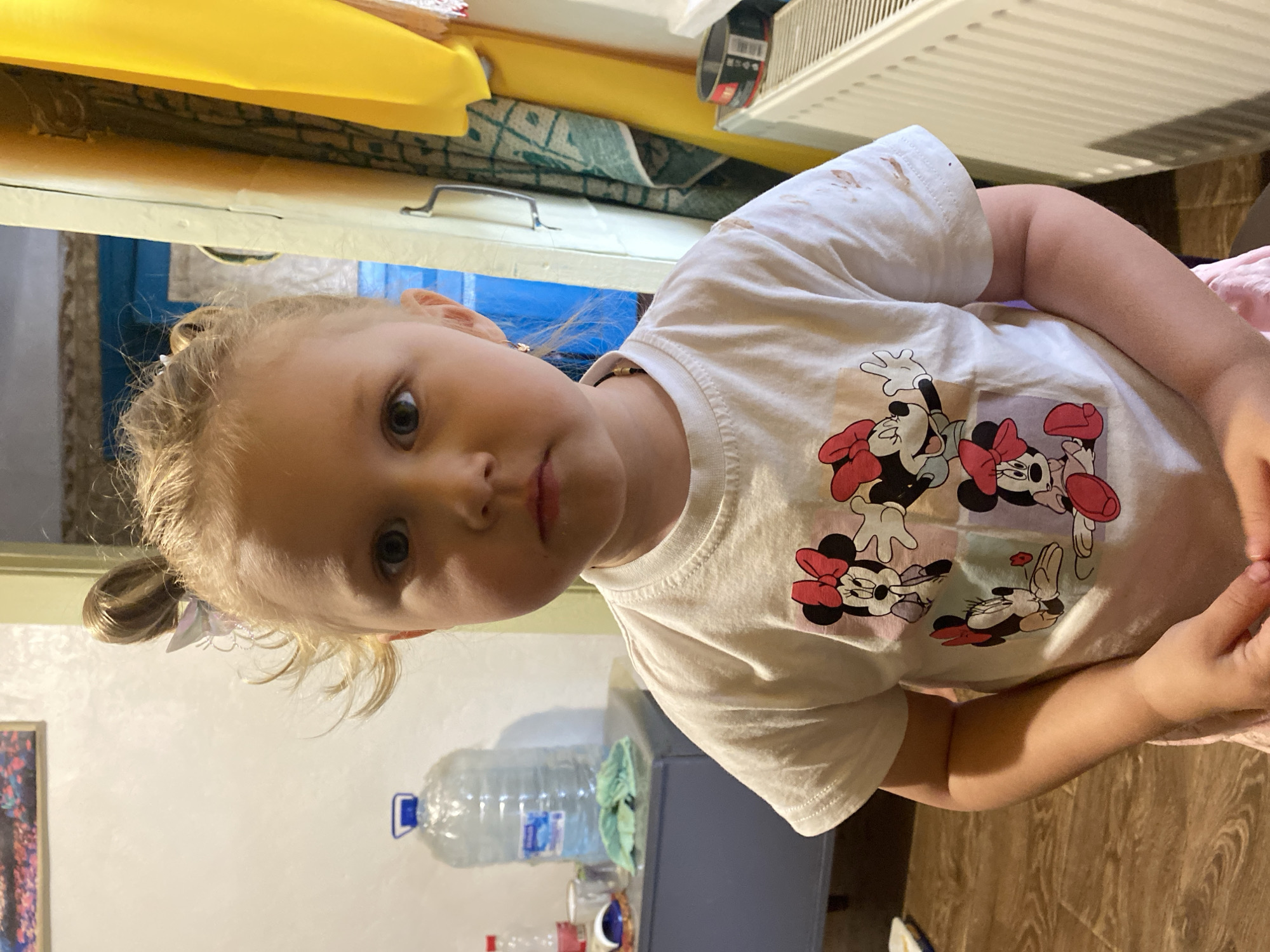 The media file shows a close-up, slightly angled portrait of a young blonde girl indoors. She is wearing a white t-shirt featuring a graphic of Minnie Mouse characters. The girl has light-colored hair pulled back loosely, with a small white bow. Her expression is neutral, and she is looking towards the camera.
Behind the girl, the background includes household items. To the left, a yellow object is visible, possibly a piece of furniture or decoration. Below that, a blue doorframe or wall section can be seen. To the right of the girl, a white ribbed radiator or vent is prominent. In the lower portion of the image, a large clear plastic bottle of water with a blue cap is present, along with other unidentifiable objects on what appears to be a table or shelf. The floor is a wooden laminate.
The lighting suggests an indoor setting, likely during the daytime, as there are no strong indications of artificial lighting. The overall scene appears to be a casual, domestic environment. No specific activity is depicted other than the girl posing for the photograph. The image is sharp and in focus on the girl's face and torso.