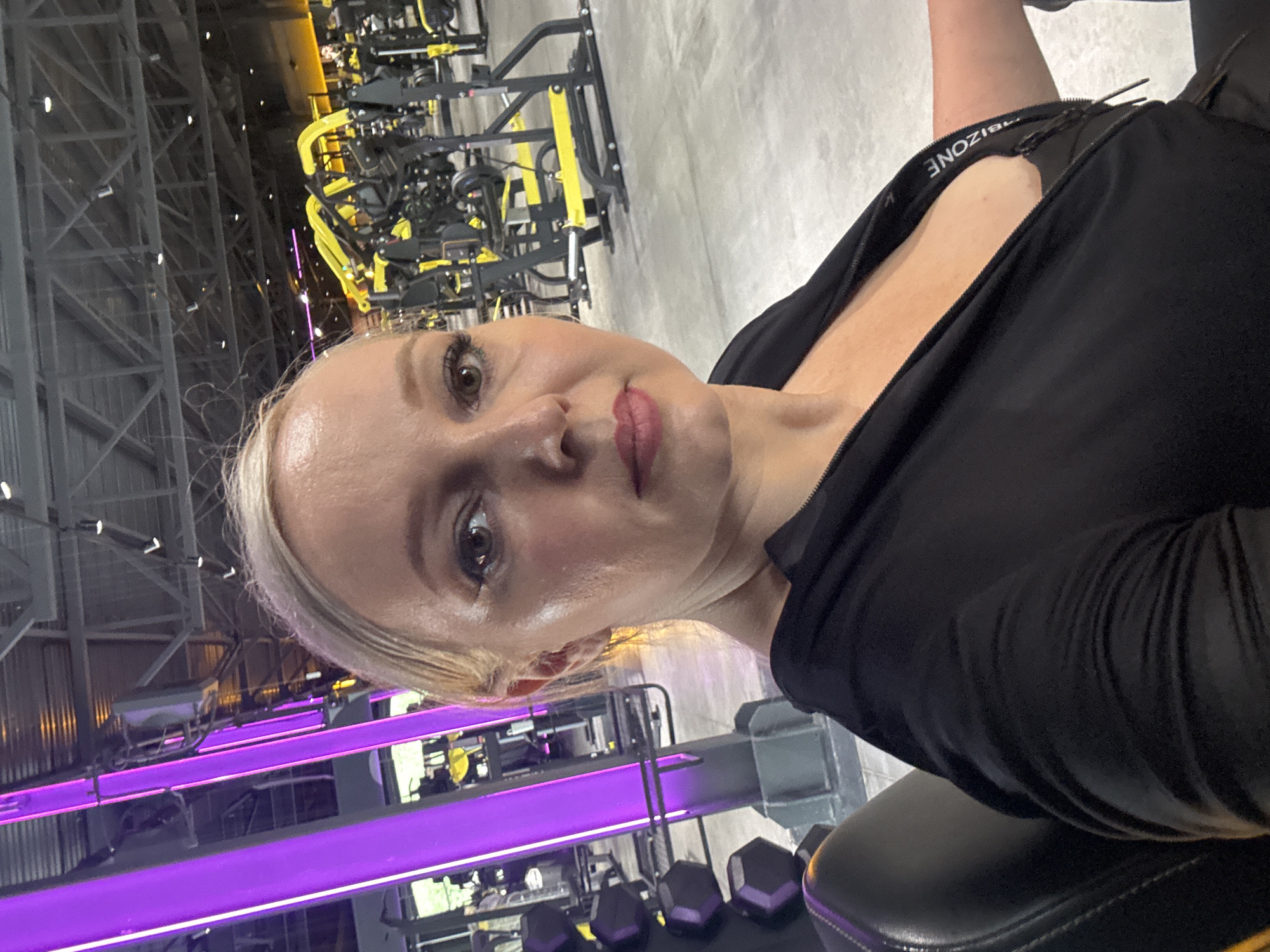 The image shows a woman in a gym. The gym is equipped with various exercise machines and weights, with a focus on yellow and black equipment. There are also bright purple lights illuminating the space, suggesting a modern and energetic atmosphere. The woman is wearing a black zippered top, partially unzipped, revealing a dark undergarment. She has blonde hair and is looking directly at the camera. The scene appears to be a personal photograph taken in a public fitness facility.