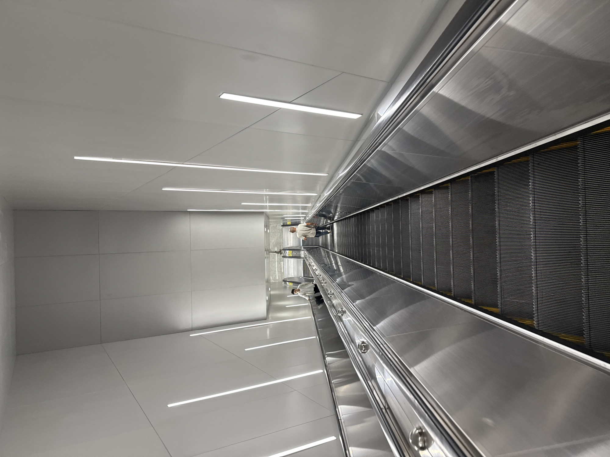 This image shows a pair of escalators in the Haizhu District, China, that are moving downwards. Two individuals are visible on the escalators. On the left escalator, a person wearing a light-colored shirt and dark pants is moving down. On the right escalator, a person in a light-colored striped shirt and dark pants is also moving down. Both escalators are constructed of polished metal and ribbed black treads. The walls of the escalator shaft are a smooth, light gray material, with vertical LED light strips illuminating the space. The ceiling is also made of the same light gray material. The overall impression is one of a modern and clean public space.