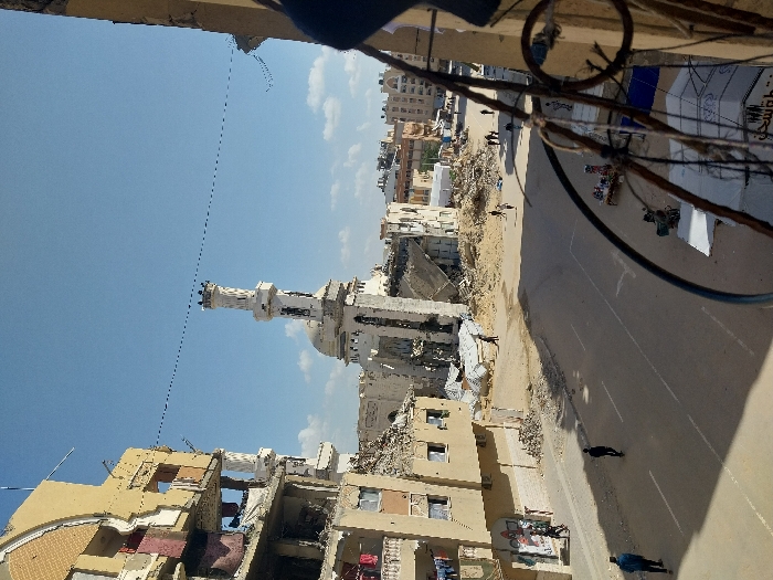 The image depicts a broad view of a street in Khan Yunis, Palestinian Territory, characterized by significant urban damage under a clear blue sky.
In the center-right, a prominent light-colored mosque, identifiable as the Great Mosque of Khan Yunis, stands with its tall, multi-tiered minaret largely intact. The main prayer hall structure, including its dome, shows extensive damage and partial collapse, with debris scattered around its base.
To the left, a multi-story building, likely residential, exhibits severe structural damage, with exposed interior rooms, collapsed sections, and visible hanging laundry. Further left, partially damaged buildings line the street. Across the street from the mosque, more damaged structures and piles of rubble are present.
The paved street, marked with white lines, runs diagonally from the left foreground towards the right background. Several individuals, primarily men, are seen walking along the street and sidewalks. On the far left, two individuals walk away from the viewer. One person walks alone in the middle of the street. Towards the right, more people are visible near the debris and makeshift structures.
In the mid-ground right, multiple tents or temporary market stalls are erected, with one displaying colorful merchandise. A white tent with Arabic writing is also visible. The foreground right is framed by a dark, ornate metal railing, suggesting the image was taken from an elevated position, such as a balcony. Overhead, thin black power or communication lines stretch across the sky. Shadows on the ground indicate a sunny day.