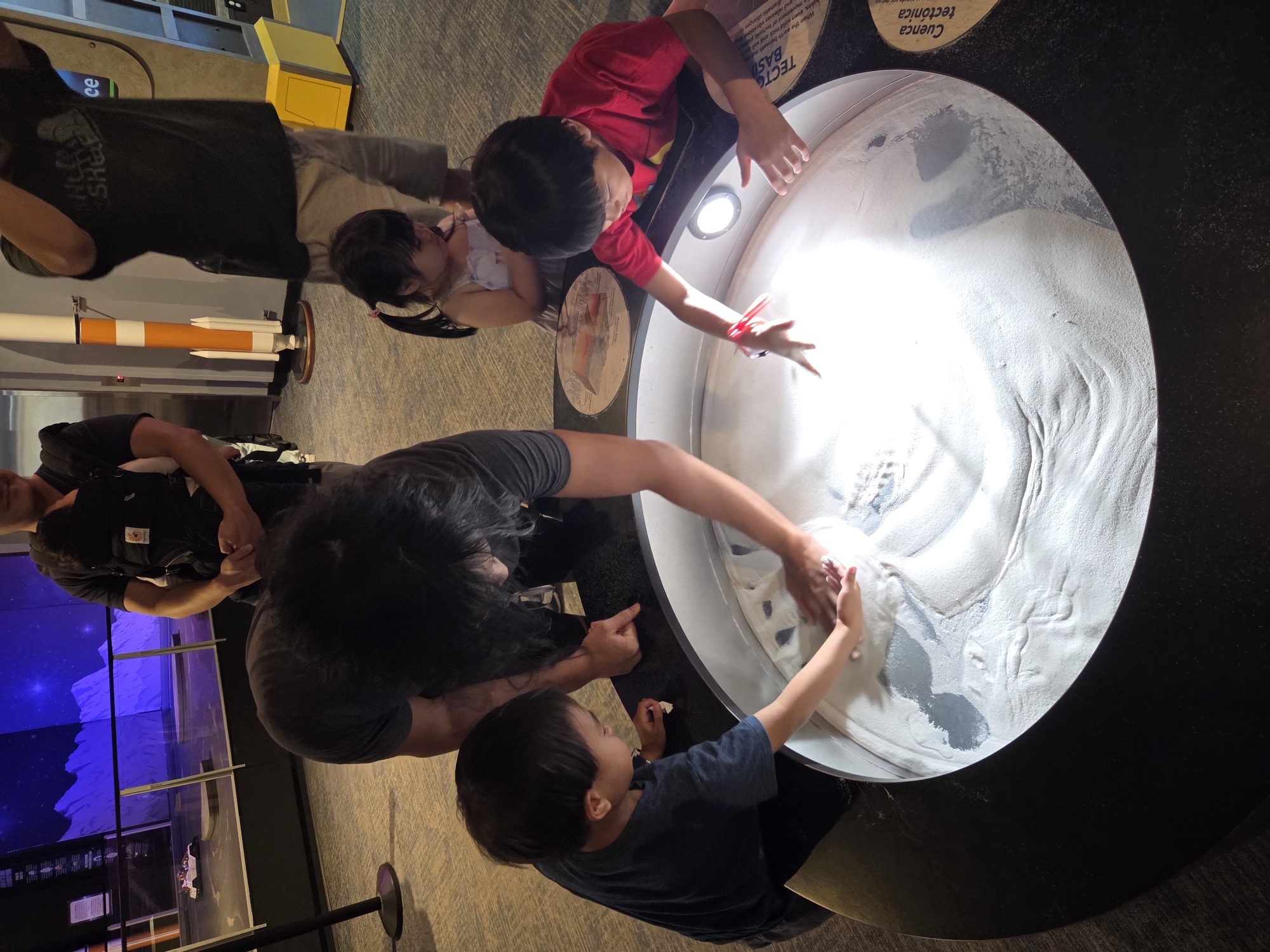 The image captures a lively scene inside what appears to be a museum exhibit. The central focus is a large, circular sand table, illuminated from below, where several children and adults are interacting with the sand. In the foreground, two young boys are engrossed in playing with the sand, with one boy on the left wearing a dark blue t-shirt and the other on the right in a red t-shirt. Both are actively using their hands to sculpt and explore the sand. An adult, with dark hair and wearing a grey t-shirt, is bent over the table, also participating in the sand play, their hands visible in the sand.
In the background, a young girl with dark hair tied in pigtails, wearing a light-colored dress, is observing the sand table. Behind her, an adult male, partially visible, is wearing a dark shirt and a carrier with a baby, suggesting a family outing. The environment surrounding the sand table features a polished floor and in the distance, a large mural or display depicting a starry night sky and lunar landscape, with a model rocket and a child-sized vehicle. The overall impression is one of educational engagement and family enjoyment within a museum setting, likely the Discovery Cube Orange County in Santa Ana, California, given its known exhibits on science and space exploration.