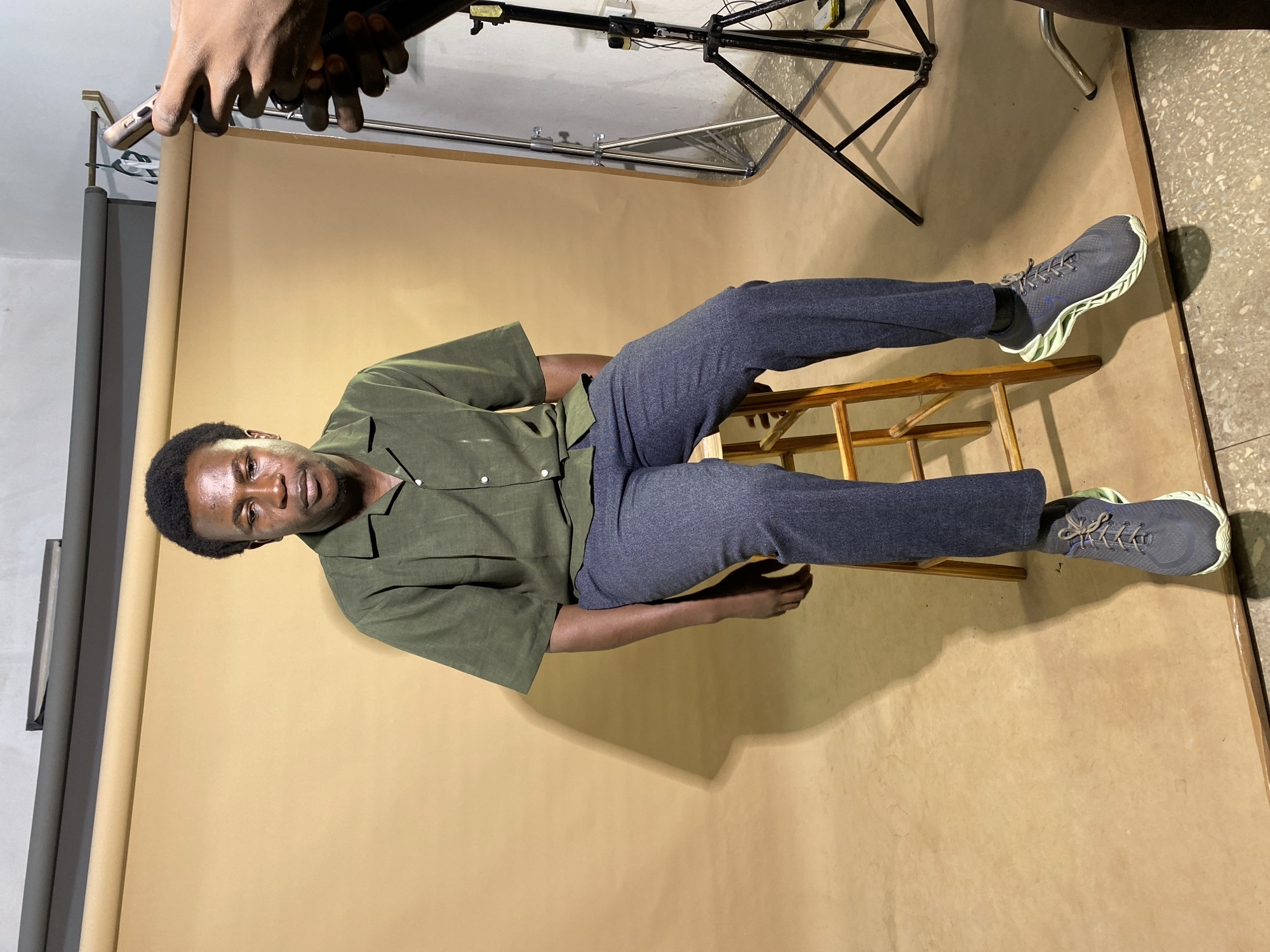 A young Black man is seated on a wooden stool in front of a tan backdrop in Gura Top, Nigeria. He is wearing a short-sleeved, olive green collared shirt tucked into dark gray trousers. His feet are clad in dark gray athletic shoes with a light green sole. To the right of the man, a hand holding a mobile phone is visible, suggesting the man is being photographed or filmed. The environment appears to be a studio setting with professional lighting equipment, including a light stand with a reflector, positioned to the right of the subject. The background consists of a seamless tan paper or fabric, typical of a photography studio, with a concrete floor visible at the bottom of the frame. Shadows are cast on the backdrop, indicating a light source from the right and slightly above.