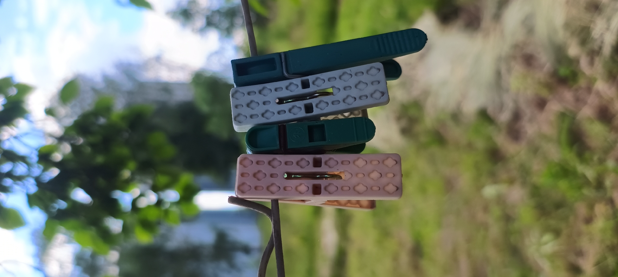 The media file displays a close-up, vertical shot of four clothespins stacked on a wire, likely a clothesline. The clothespins are of varying colors: the top one is dark green, followed by a white one, then another dark green one, and finally a light pink or beige one at the bottom. The white and pink clothespins have an embossed pattern of four petal-like shapes with a circle in the center. The green clothespins have a ribbed texture on the part that grips. They are all positioned horizontally and appear to be tightly clipped onto the wire.
The background is out of focus, suggesting a shallow depth of field. It shows a blur of green foliage, indicative of trees or bushes, and a bright, hazy sky with hints of blue and white. The overall impression is an outdoor setting, possibly a backyard or garden, on a bright, likely sunny day, given the brightness of the sky. There are no people or animals visible in the frame. The image is static, with no discernible activity or event taking place beyond the clothespins being attached to the wire. The lighting suggests it could be daytime, though the exact time is not specified. There is no visible text in the image. The focus is entirely on the arrangement and details of the clothespins.