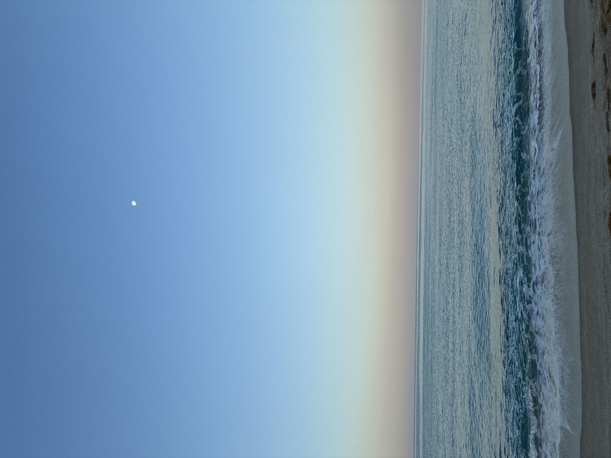 A photograph captures a serene seascape with a clear sky and gentle waves lapping at a sandy beach. The sky transitions from a deep blue at the top to a soft gradient of pale blue and warm peach hues near the horizon, hinting at either dawn or dusk. A small, pale moon is visible in the upper portion of the sky. The ocean is a tranquil turquoise, with white foam cresting on the waves as they approach the shore. The beach in the foreground is composed of fine sand, with a few faint footprints visible. The scene is devoid of any people or structures, emphasizing the natural beauty of the location. This scene could be representative of a quiet beach in Alanya, Turkey, during the early morning or late evening.