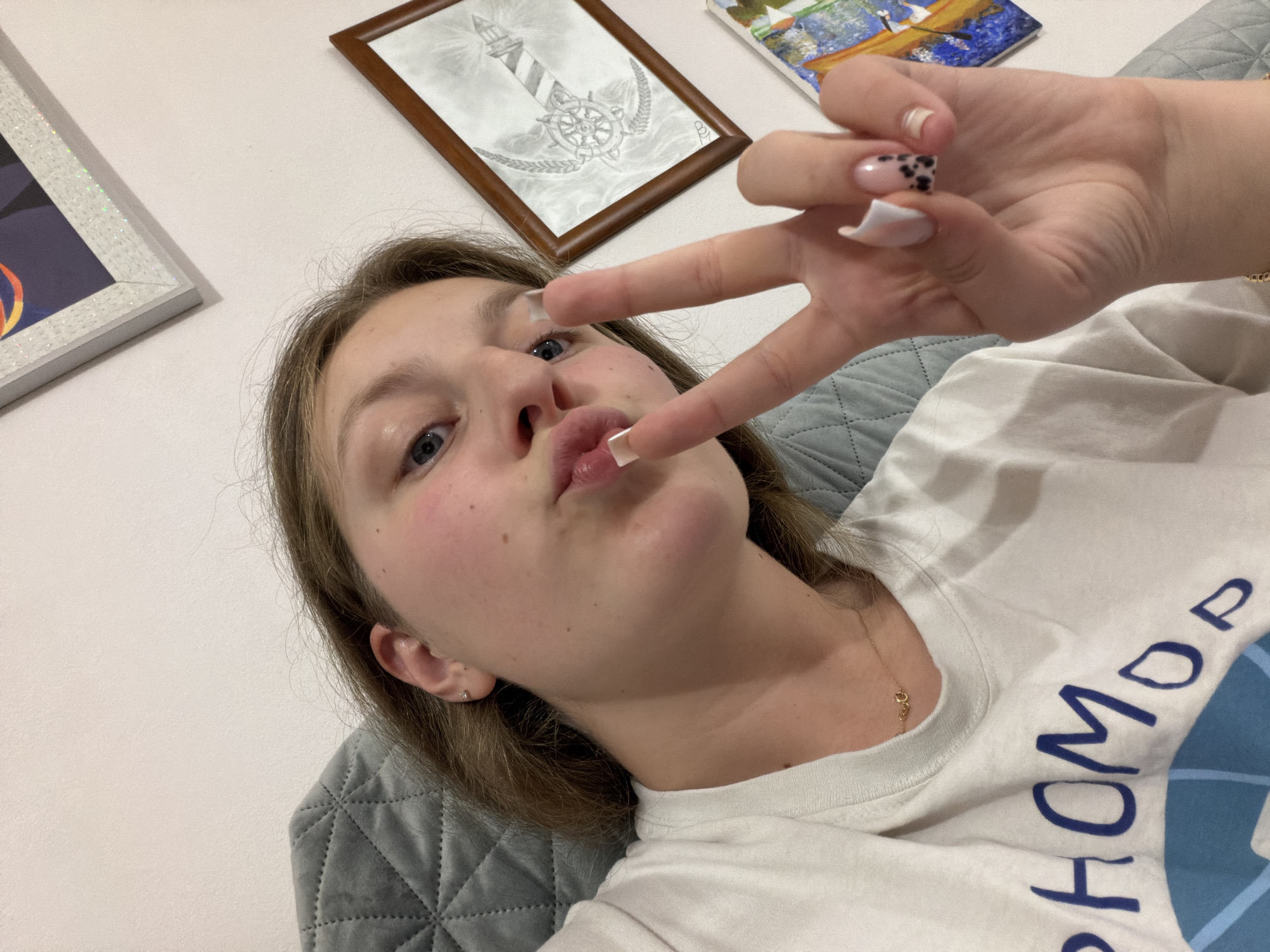 A young woman, with light brown hair and blue eyes, is shown in a close-up, slightly angled selfie. She is wearing a white t-shirt with a blue graphic and text, a delicate gold necklace, and has a small stud earring in her left ear. Her lips are pursed as she makes a peace sign with her right hand, which features manicured nails with a black and white spotted design. She appears to be indoors, seated on a gray quilted surface, possibly a chair or sofa. The background includes a plain white wall adorned with two framed pictures. One frame is ornate and white with glittery accents, displaying a colorful abstract image. The other, a smaller, wooden-framed picture, contains a detailed pencil drawing of a nautical helm and lighthouse. The scene is lit by artificial light, creating soft shadows. The overall impression is casual and personal. The location is not specified in the image, therefore it cannot be determined if it is in Burgas, Bulgaria.
