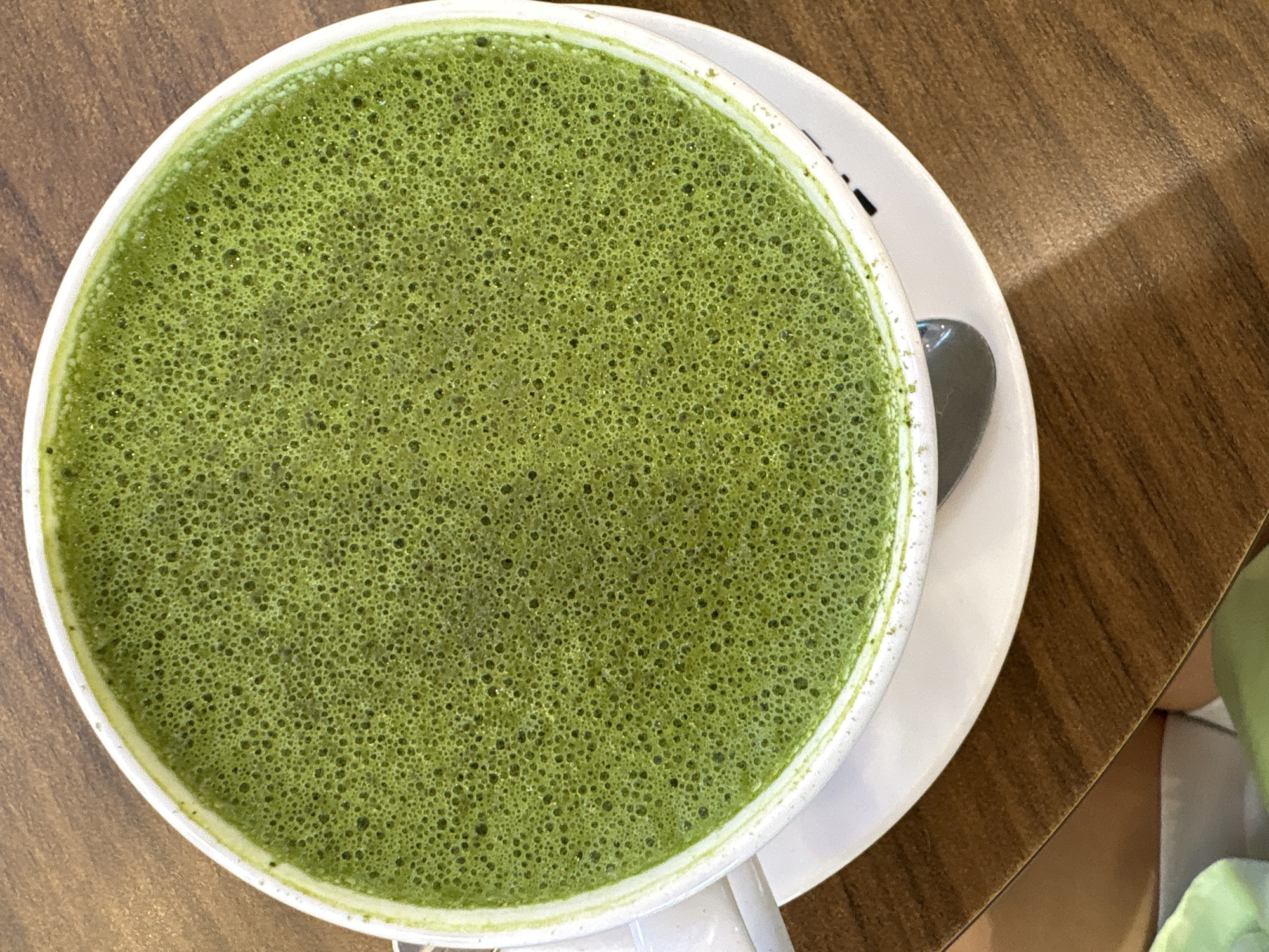 An aerial shot reveals a cup of vibrant green matcha latte, characterized by a frothy surface dotted with tiny bubbles. The cup sits on a white saucer alongside a small spoon, positioned on a wooden table. A glimpse of an individual and a green bag is visible in the periphery. While the image suggests a café setting, the specific location in Nha Trang, Vietnam, cannot be determined without additional contextual information.