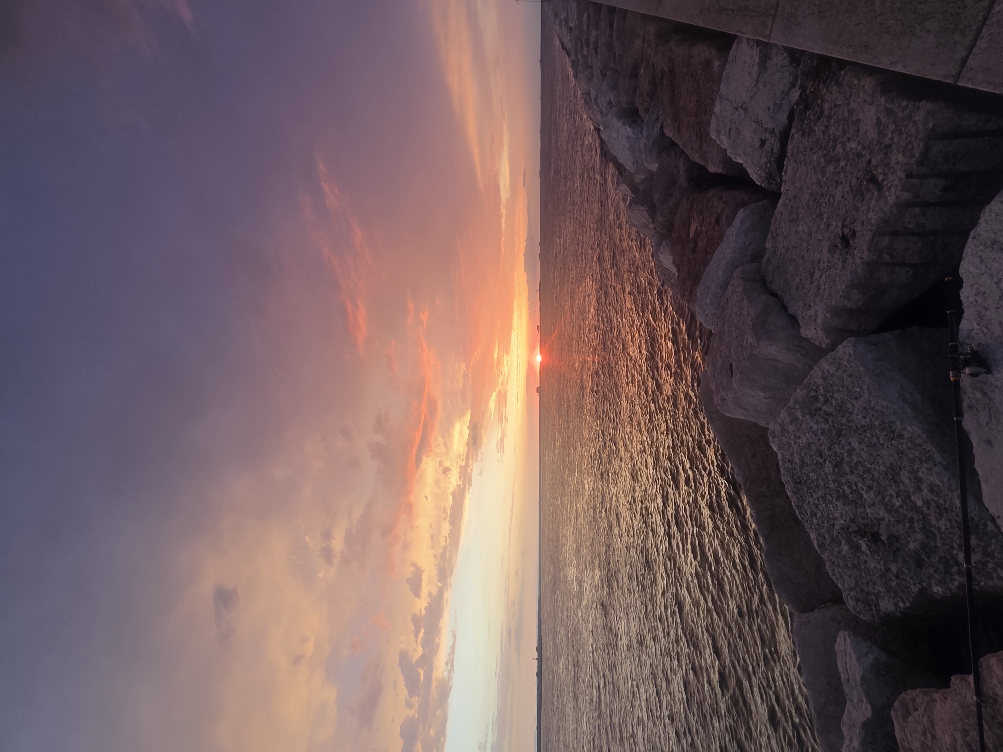 The image depicts a sunset over the Adriatic Sea, viewed from a breakwater composed of large, irregular grey stones. The sky is a gradient of vibrant colors, transitioning from a soft orange and pink near the horizon to a deep purple at the top. Clouds are scattered across the sky, catching the sunset's light. The sea is calm, with gentle ripples reflecting the warm hues of the sky. In the distance, several ships and what appears to be a lighthouse are visible on the horizon. The breakwater extends to the right of the frame, with a paved walkway visible on its far edge. Two fishing rods are positioned on the rocks in the foreground. The scene suggests a peaceful evening by the water, likely in the city of Chioggia, Italy, known for its coastal setting and maritime activities.
