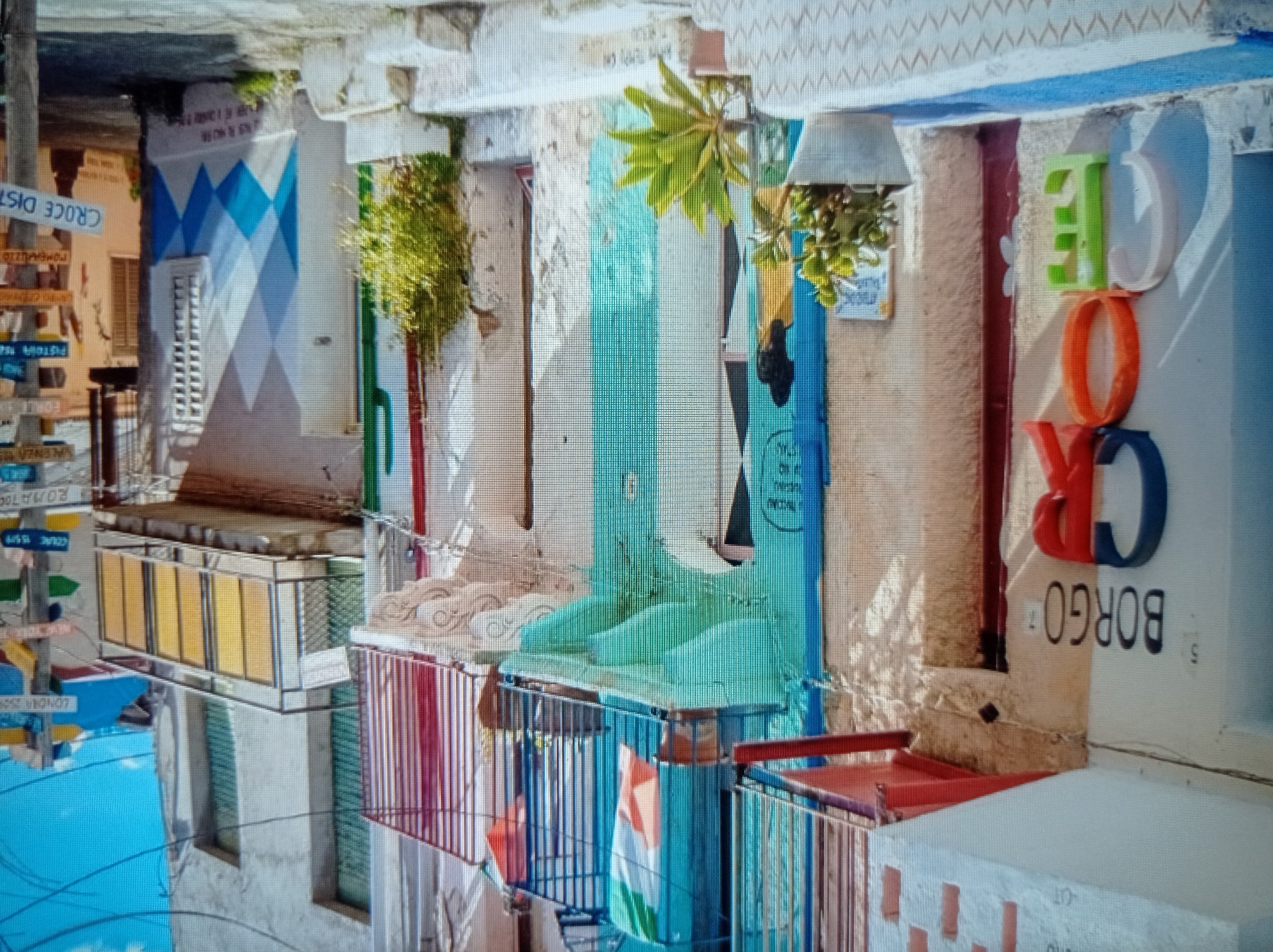 The image depicts a narrow street scene in what appears to be a European town. The buildings are multi-story and painted in various colors, with balconies adorned with railings. An Italian flag is visible hanging from a balcony, suggesting a connection to Italy. Several street signs are present, pointing to different locations, including "LONDON 2520", "NEW YORK", and "ROMA". The sign "CROCE DIST" is also prominently displayed, which translates to "Cross Distant" in Italian. Large, three-dimensional letters spelling "BORGO CROCE" are mounted on one of the building's facades. Potted plants are placed on steps leading up to the buildings, adding a touch of greenery to the scene. The architecture and street signs suggest a vibrant, possibly coastal or historically significant, urban environment. The overall impression is one of a picturesque and lively street, likely in a Mediterranean locale, but it is not located in Mukachevo, Ukraine.