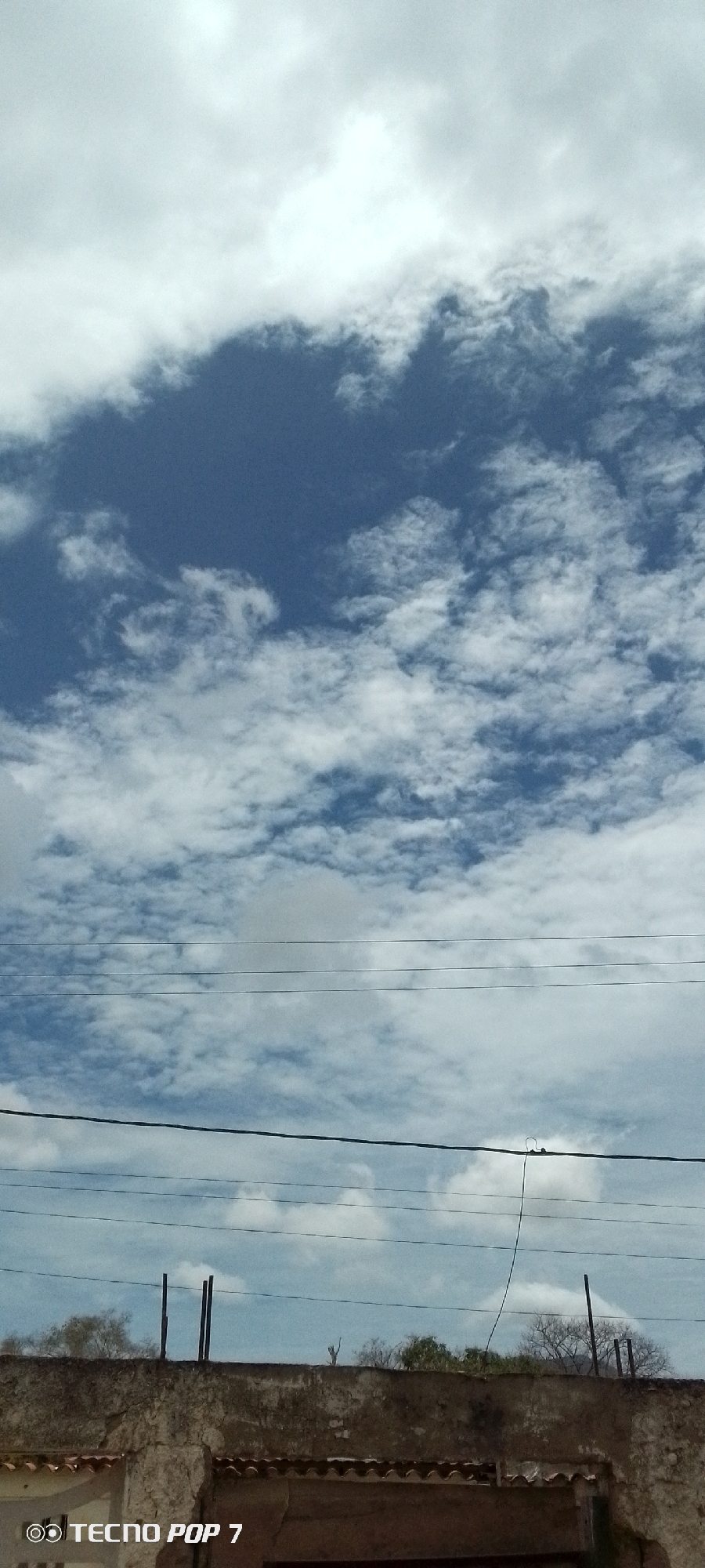 The image shows a partly cloudy sky in Altagracia, Venezuela. In the foreground is a weathered, stone or concrete structure. Two dark posts are extending from this structure. Behind the structure are treetops, visible beyond. There are several electrical or telephone lines in the middle ground, running horizontally across the image. The sky above is a mix of blue and white clouds. The lower left corner of the image includes a "TECNO POP 7" watermark.