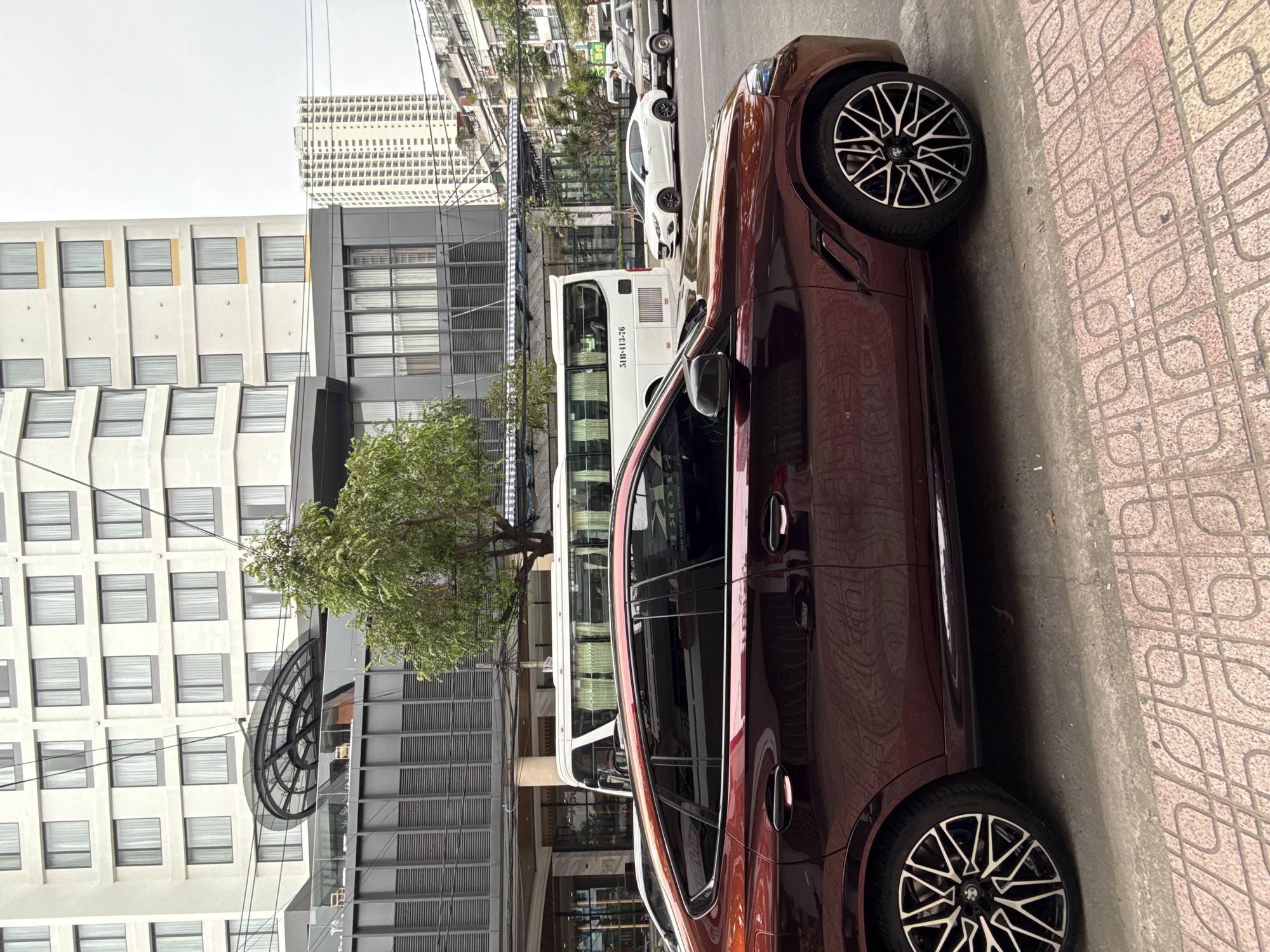 The image depicts a brown vehicle parked on a sidewalk in Nha Trang Ward, Vietnam. The sidewalk is patterned with interlocking red and gray designs. Adjacent to the sidewalk, a multi-story building with rows of rectangular windows stands tall, along with a tree and an architectural element resembling a circular canopy. Further into the scene, a white bus and a white car are visible on the street, with additional buildings and infrastructure extending into the background. Overhead power lines crisscross the sky.
