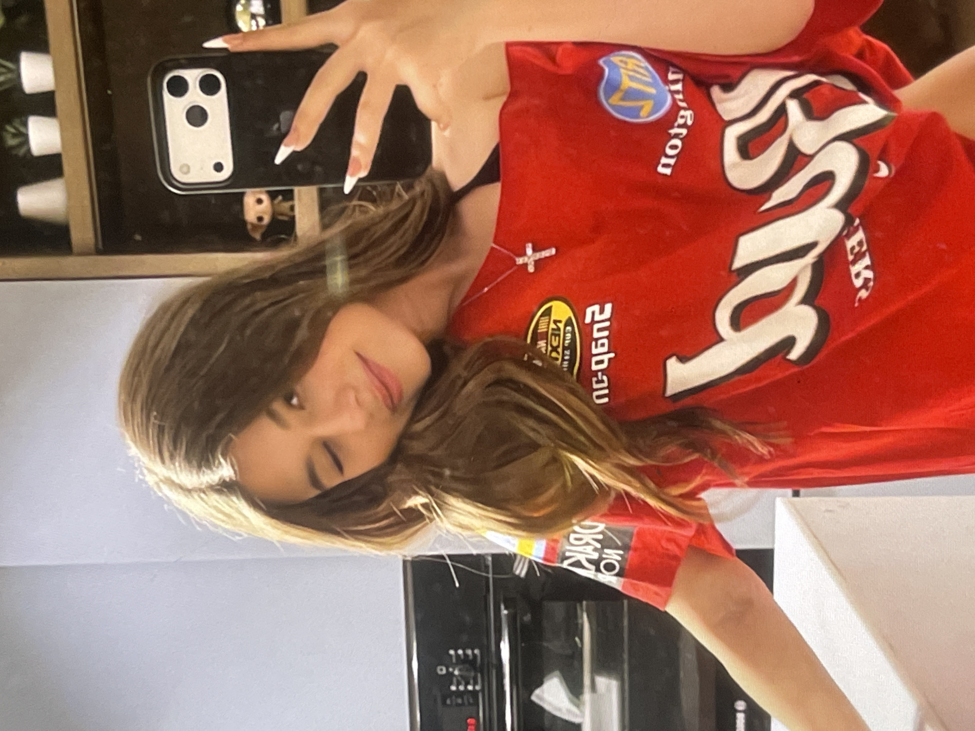A young woman is captured in a mirror selfie, holding a black smartphone with a triple-lens camera module. The image is rotated 90 degrees counter-clockwise. She has long, light brown hair, and is wearing a red t-shirt adorned with various graphics and text. A silver cross necklace is visible around her neck. She has long nails with white tips. Her face shows a slight smile and one eye is closed in a playful wink.
The red t-shirt features stylized white and black text, which is mirrored due to the selfie. Partially decipherable text includes what appears to be "JAGGER" or similar in large white font, and patches with "NEXUS," "MON," and a blue and yellow patch reading "BILL" (all unmirrored interpretations).
In the background reflection visible through the phone, there is a small figure, possibly a Funko Pop doll, resting on a wooden shelf with several small white pots or cups. The overall setting appears to be indoors, likely a domestic space like a kitchen or dining area, with part of a dark appliance and light-colored walls also visible in the reflection. The scene evokes a casual, everyday moment in Poltava, Ukraine.