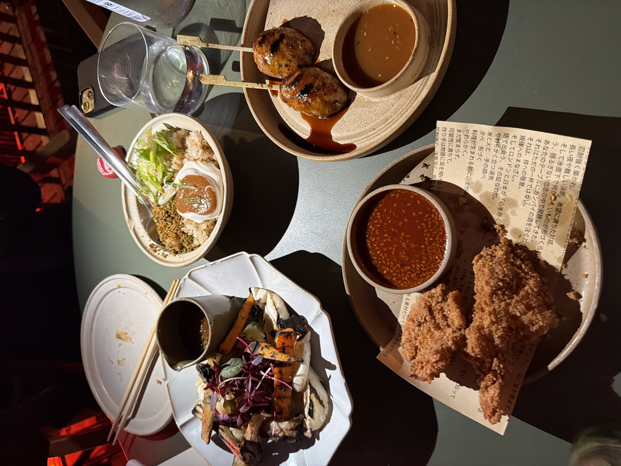 An overhead, close-up shot captures a table set for a meal, likely in a restaurant. In the foreground, two pieces of crispy fried chicken are presented on a light brown, circular plate, accompanied by a small bowl of reddish-orange dipping sauce. To the right, another light brown plate holds two grilled meatballs on skewers, with a small bowl of a lighter brown sauce. Behind these, a bowl of rice with green onions and what appears to be a dollop of white sauce or yogurt, and a small pitcher of sauce are visible. A glass of water sits to the right of the rice bowl. In the background, an empty white plate with chopsticks rests on it, suggesting the meal is in progress or nearing completion. The table surface is a muted green, and the background is dimly lit with hints of red lighting. The image does not provide any explicit visual cues to pinpoint a specific location within London, United Kingdom.