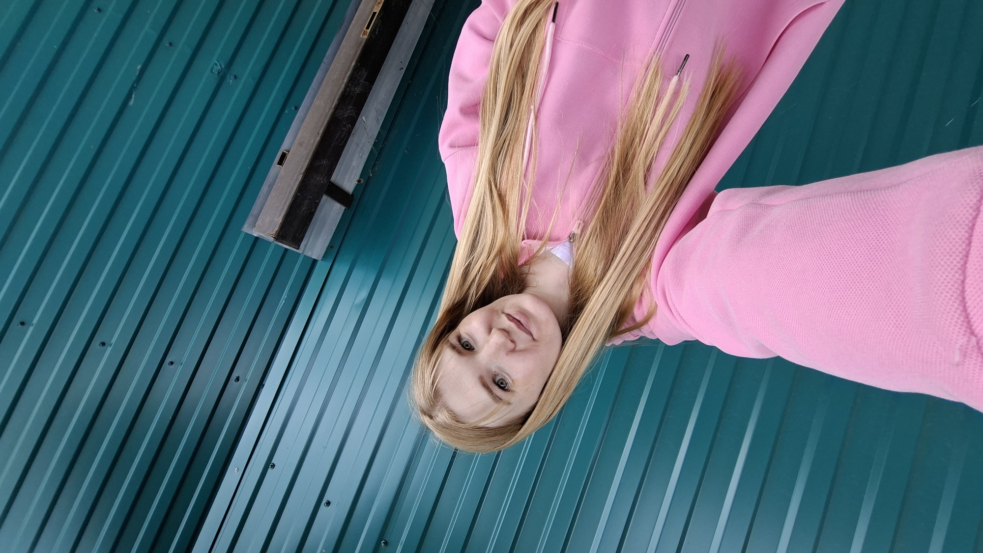 A young woman with long blonde hair and fair skin is taking a selfie in front of a teal-colored corrugated metal wall. She is wearing a pink hooded sweatshirt. To the right of the woman, there is a dark wooden beam attached to the wall. The scene appears to be outdoors, possibly in an industrial or utility area, given the materials of the wall and the presence of the wooden beam. The lighting suggests it is daytime. The location is specified as Bolshoy Bukor, Russia.