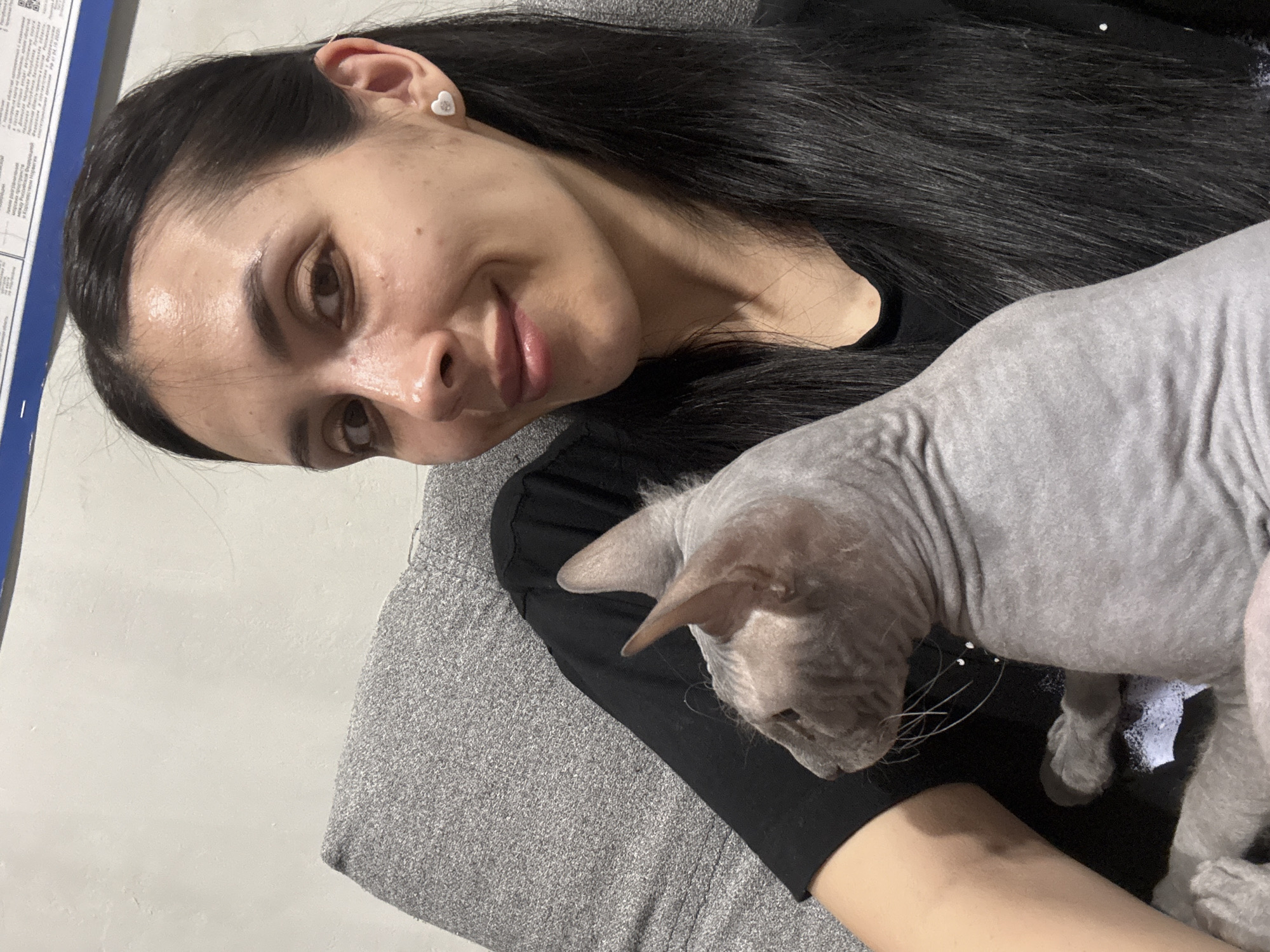 A woman with dark hair and a light complexion is pictured alongside a hairless cat, likely a Sphynx breed. The woman is wearing a black top and heart-shaped earrings. She is holding the cat against a grey, textured surface, probably a piece of furniture. The background is a plain wall, partially obscured by a blue and white sign. There is no information to indicate that this image was taken in Votkinsk, Russia.
