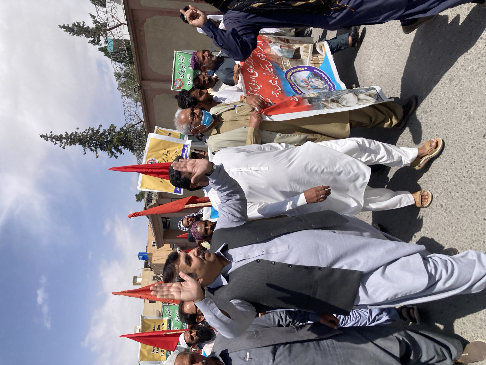 A group of men is gathered outdoors on a bright, sunny day, participating in what appears to be a public demonstration or rally. Most are wearing traditional shalwar kameez, with some sporting waistcoats. Several individuals are gesturing with raised hands, suggesting engagement in an address or chanting.
They are holding numerous banners and red flags. The banners display text predominantly in Urdu (and possibly Pashto), along with some English. Visible text includes "کیسکو لیبر یونین" (KESC Labor Union), "زنده باد زنده باد" (Long live, Long live), and parts of slogans like "در سر کوشل سیکیوں میں رجہ". English text on banners includes "OBHATA HIGH", "BMWF * ONERS FEDERATION", "Gov", "stan", "the 176", "ILO", and "PU", likely referencing government, specific locations or organizations (like Balochistan or Pakistan, and the International Labour Organization), and possibly a specific provincial government or labor law number. One banner also mentions "مائنز ایندپهنه گشت ورک" (Mines and related work).
The setting features an asphalt ground, with a building and a fence topped with barbed wire visible in the background. Several pine trees are also present behind the fence. The clear blue sky with sparse white clouds, coupled with strong shadows, indicates it is daytime, likely mid-morning to early afternoon. The scene takes place in an urban or semi-urban area, consistent with the provided location context of Quetta City Tehsil, Pakistan.