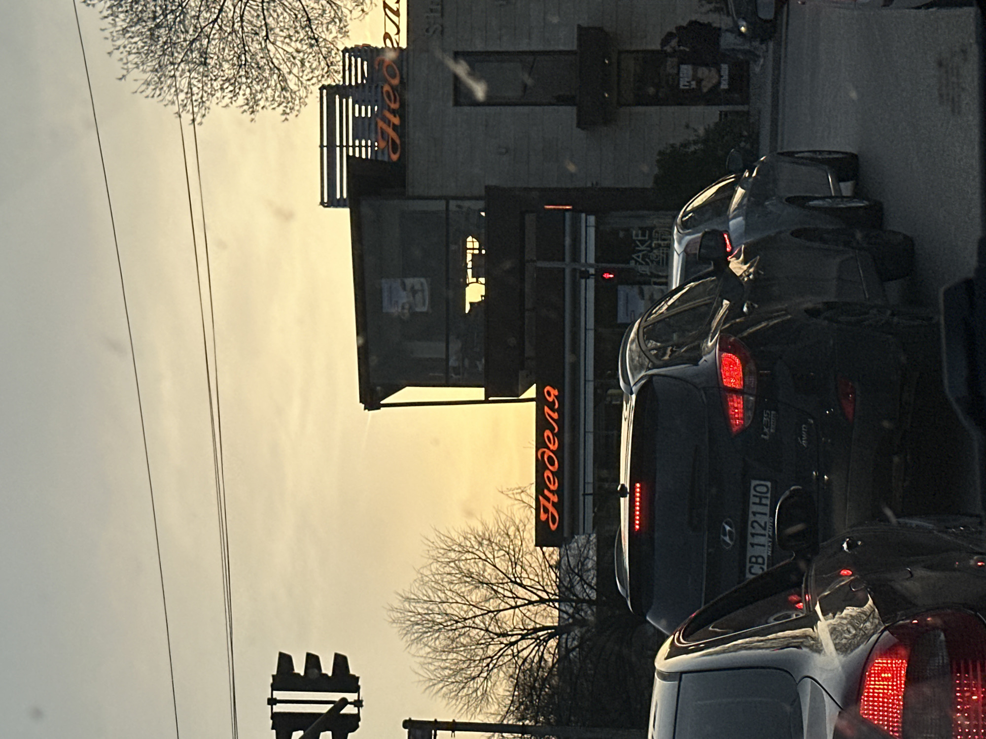 The image depicts a street scene in Sofia, Bulgaria, likely taken from within a car stopped in traffic. Prominently visible is the "Nedelya" cake shop building, with its name displayed in orange Cyrillic script. A traffic light is visible on the left. Several cars are in view, their brake lights illuminated. The sky is overcast, suggesting either early morning or late afternoon. Bare branches of trees are seen between the building and the sky. The license plate "CB 1121 HO" is visible on a dark SUV. The overall atmosphere suggests a typical urban setting during a time of day with subdued lighting.
